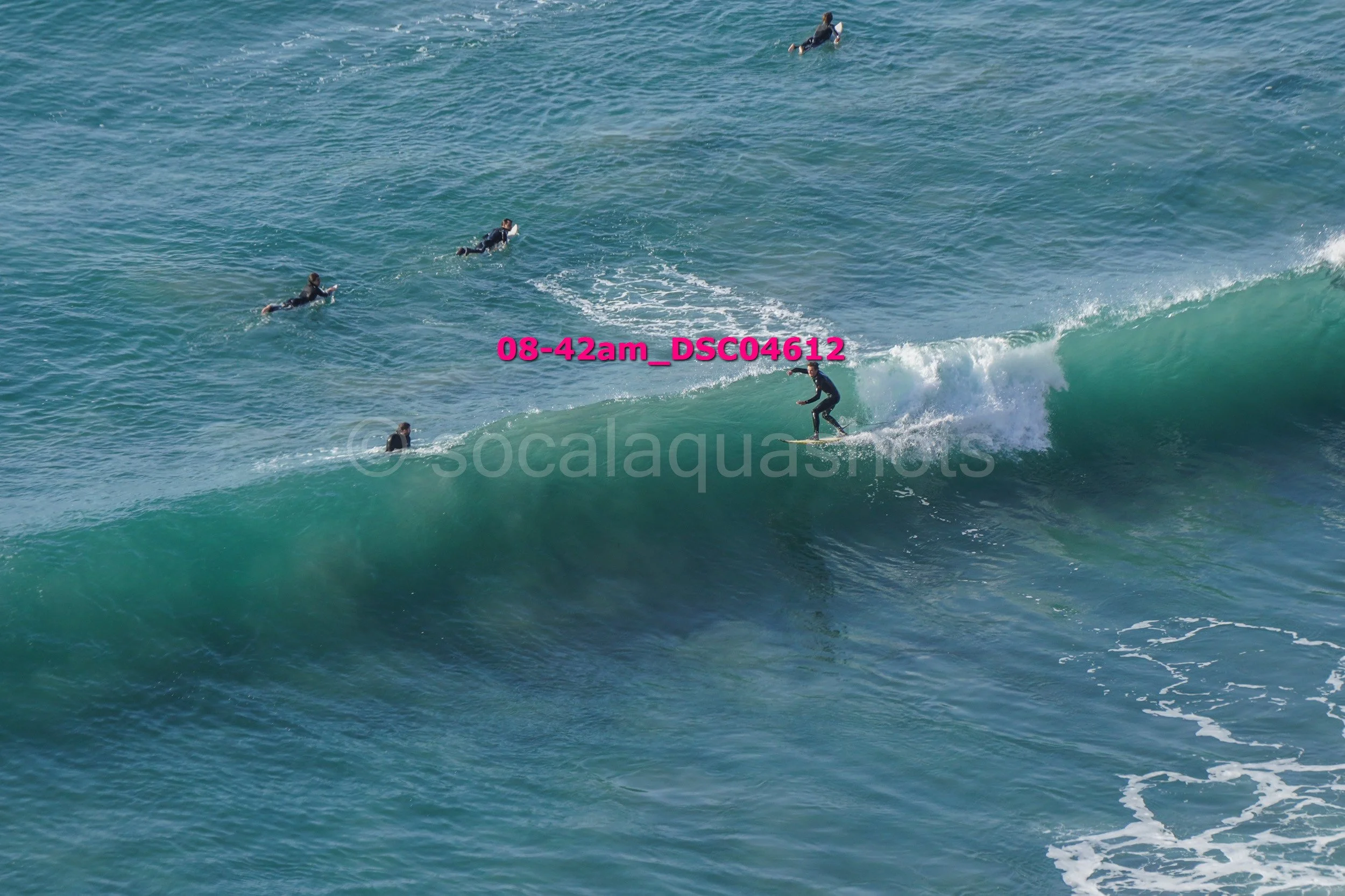 Surfer riding a wave with several people in wetsuits swimming or floating nearby in the ocean.
