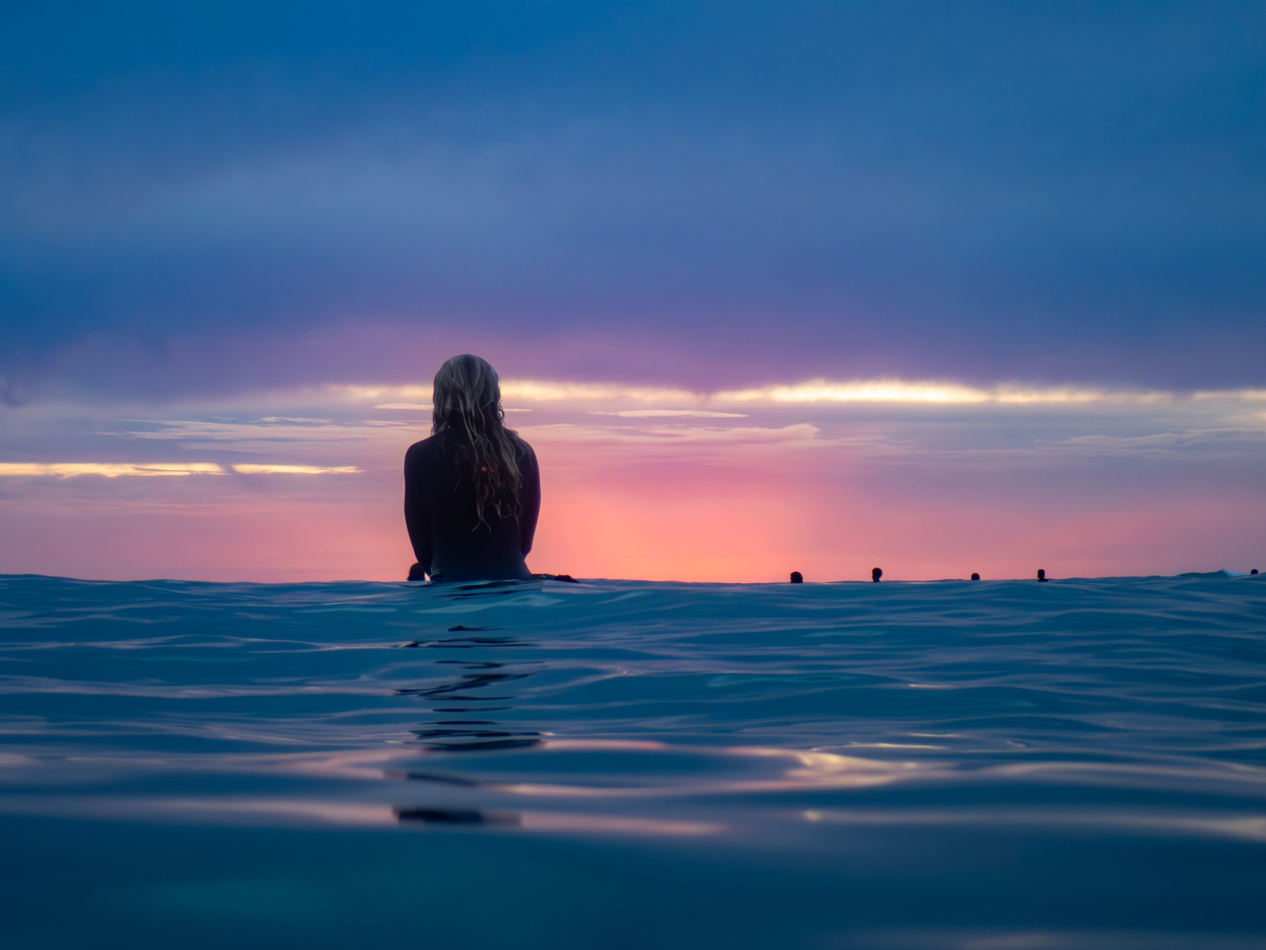 A person with long hair sitting in the ocean during sunset, facing the horizon with a colorful sky of purple, pink, and orange hues.
