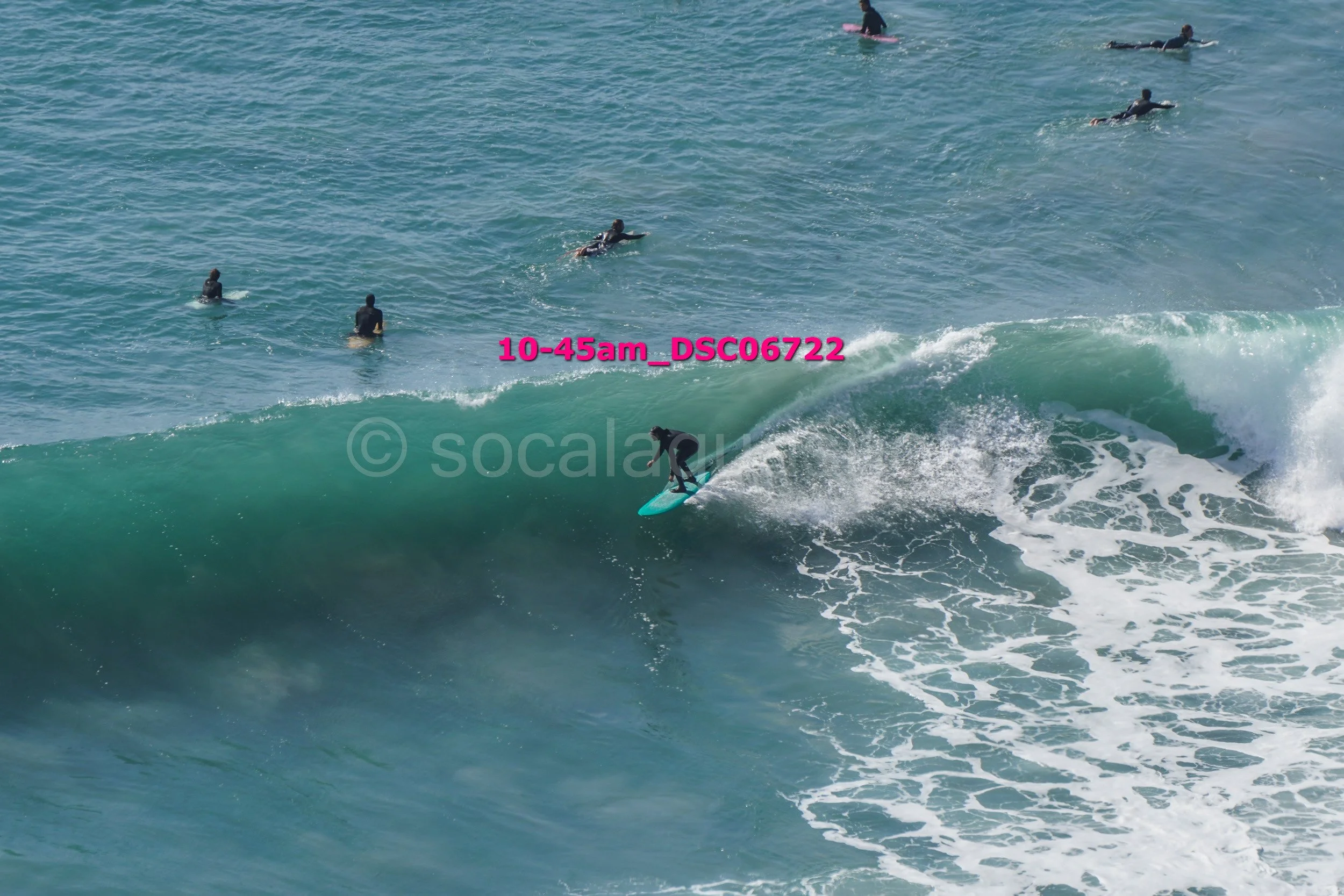 A surfer wearing a black wetsuit riding a large green wave with several other surfers in the water nearby.
