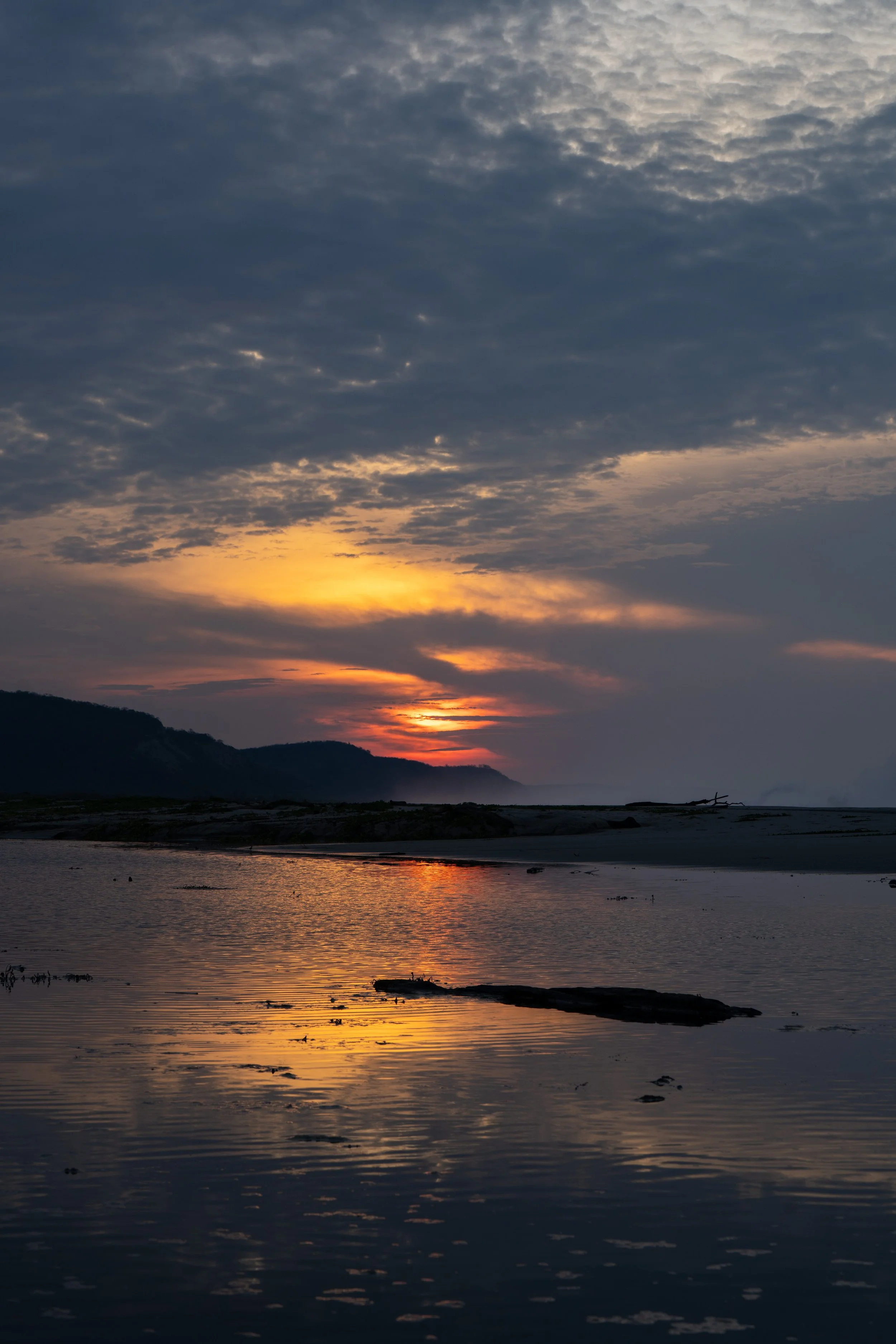 Sunset over a body of water with mountains in the background, clouds in the sky, and reflections in the water.