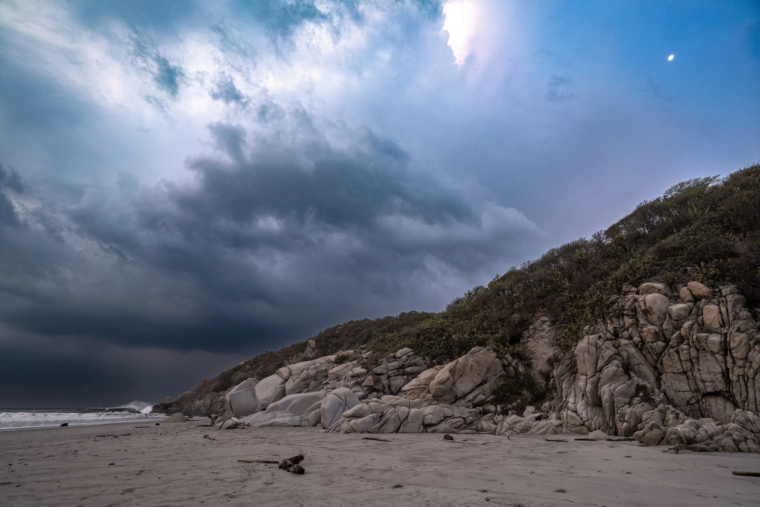 Cloudy sky over a rocky cliffside on a beach, with some waves and driftwood on the shore.