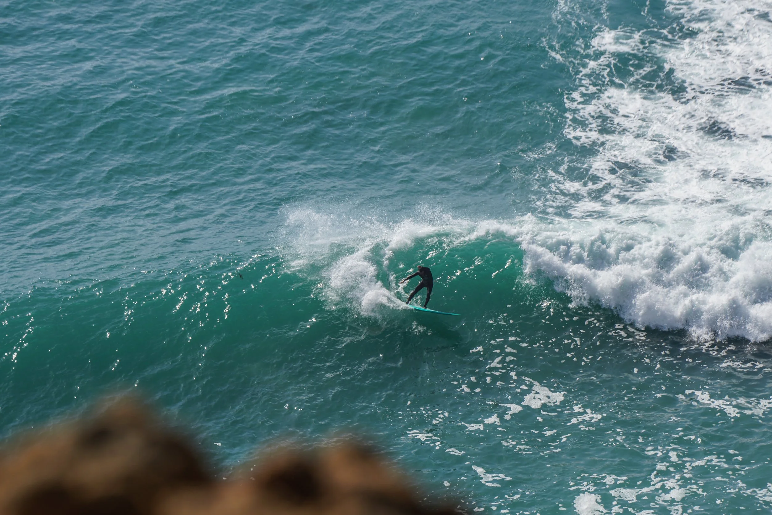 A person surfing on a wave in the ocean with a rocky shoreline in the foreground.