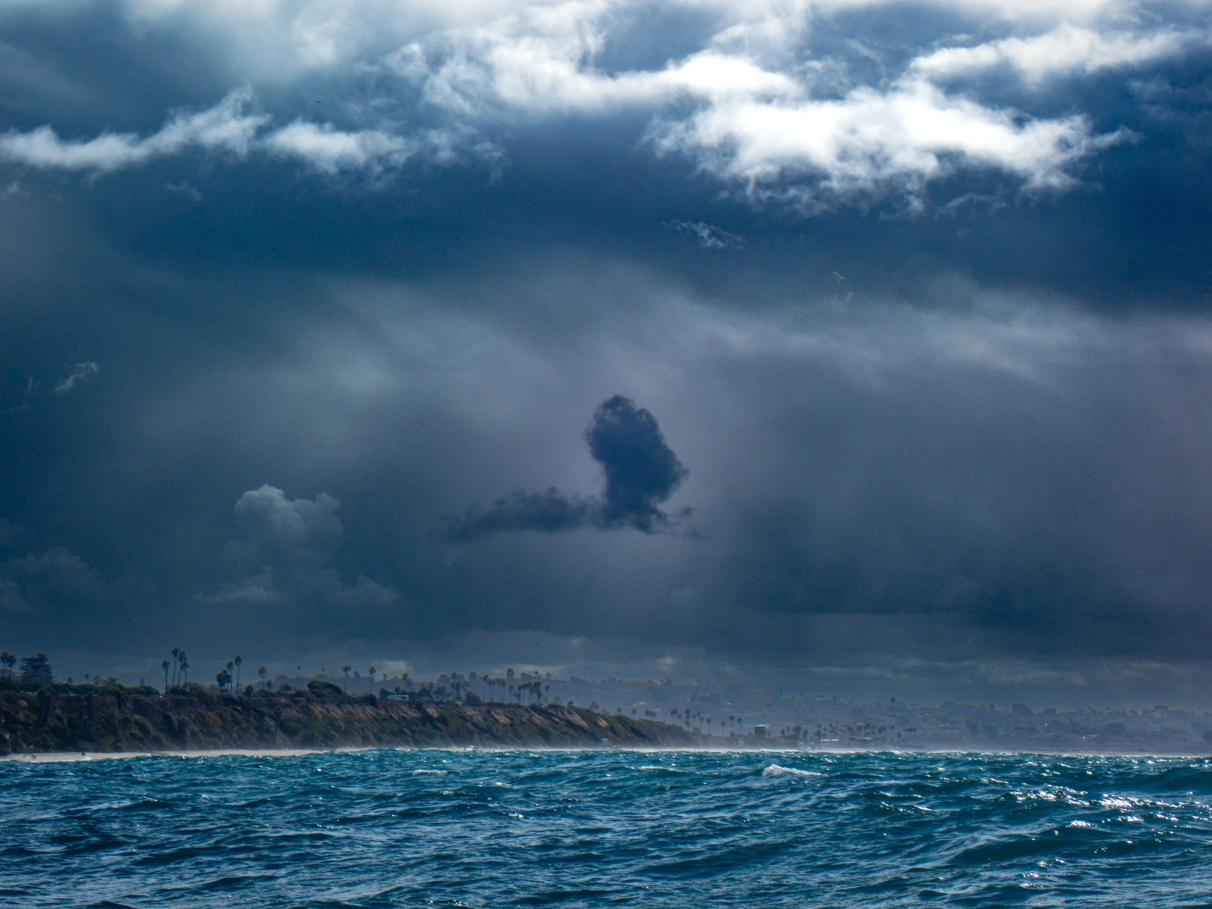 Overcast sky with dark clouds over the ocean and a distant shoreline with palm trees.