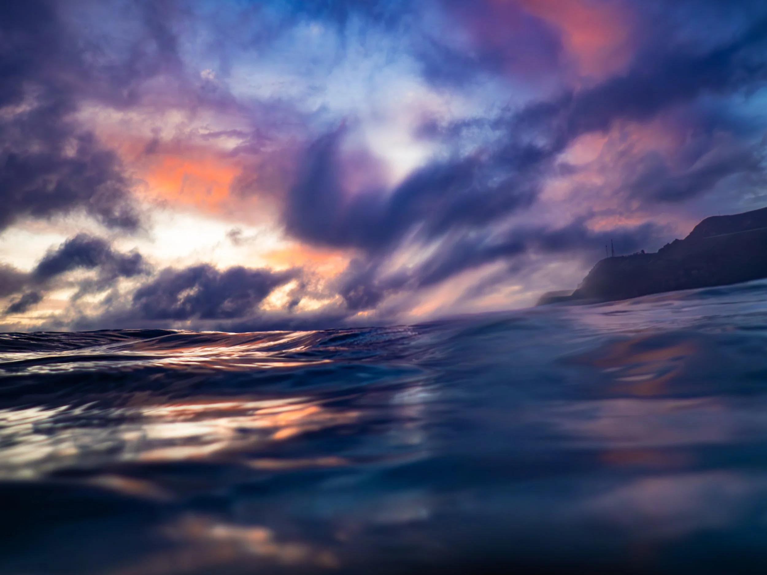 Ocean water with a colorful sunset sky filled with dark clouds and a mountainous coastline in the background.
