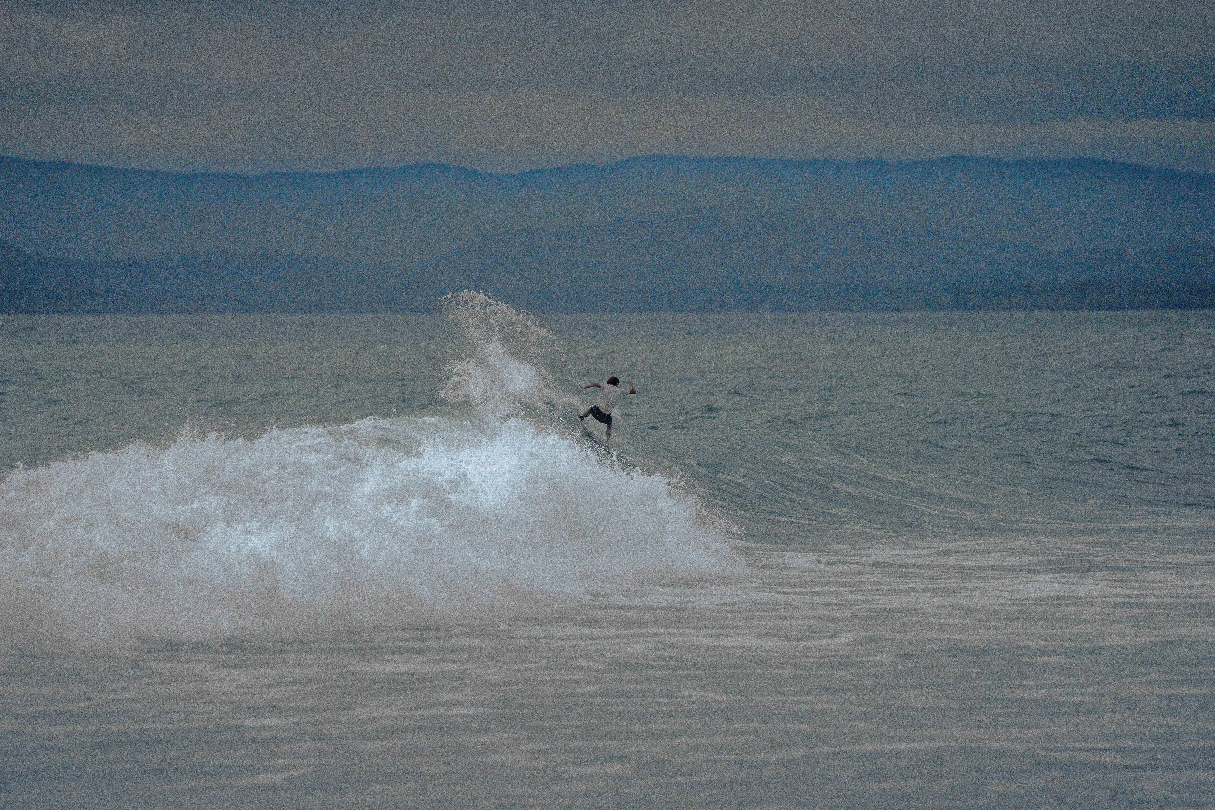 A person riding a wave on a surfboard in the ocean with mountains in the background.