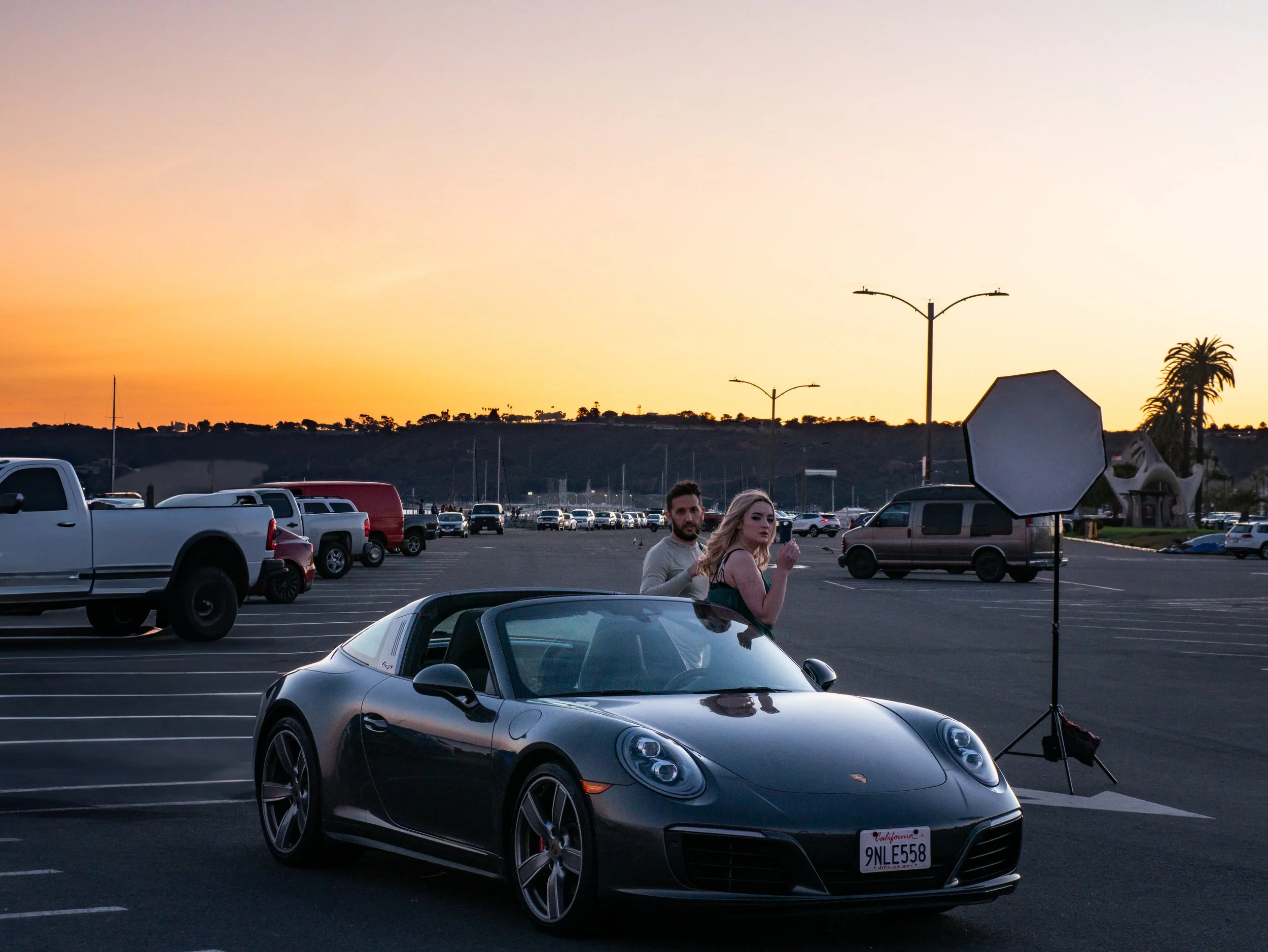 Two people, a man and a woman, standing next to a black Porsche convertible in a parking lot at sunset, with photography equipment set up nearby.