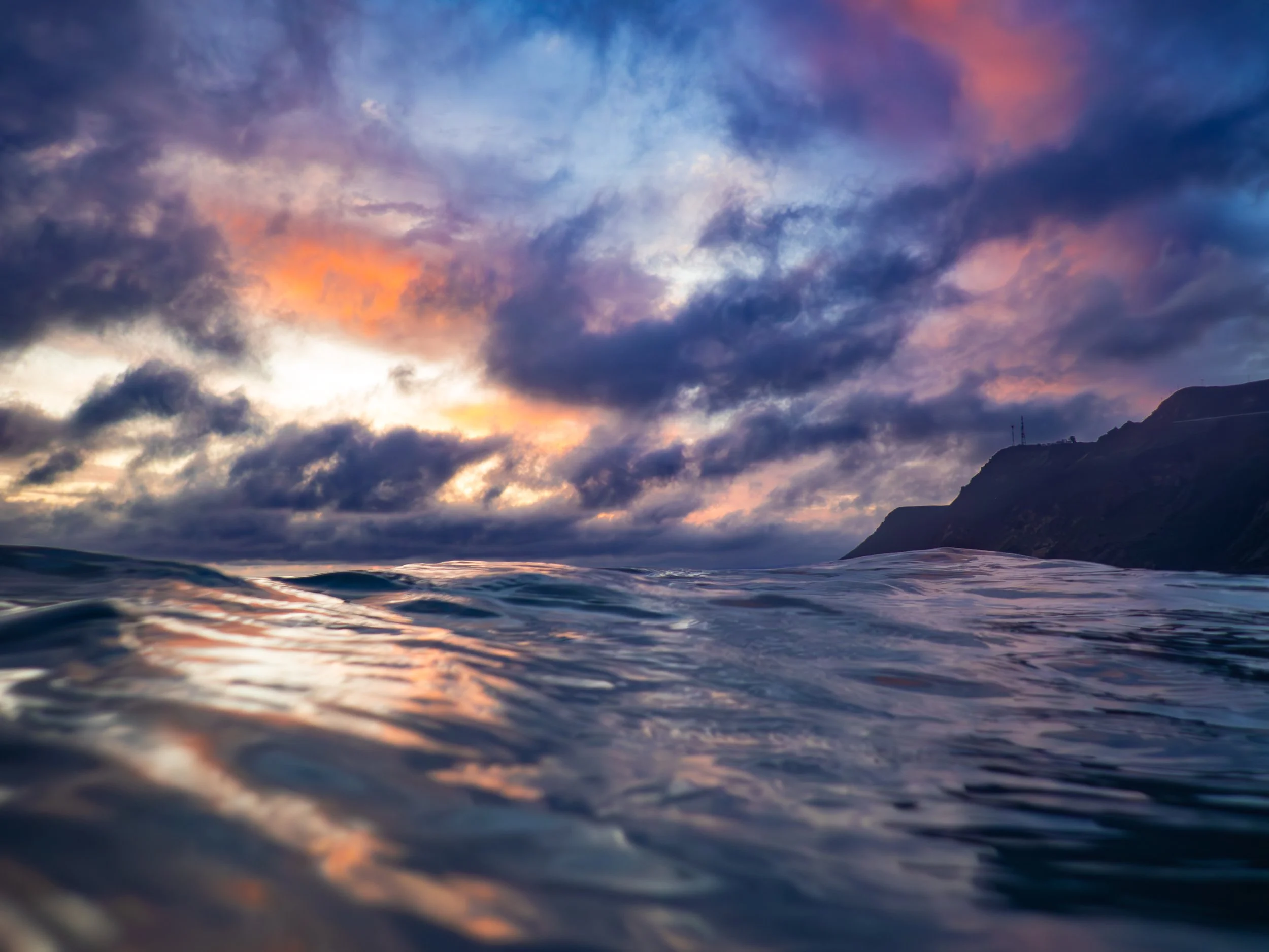 A view of the ocean with small waves, a cloudy sky with orange and purple hues at sunset, and distant cliffs on the right side.