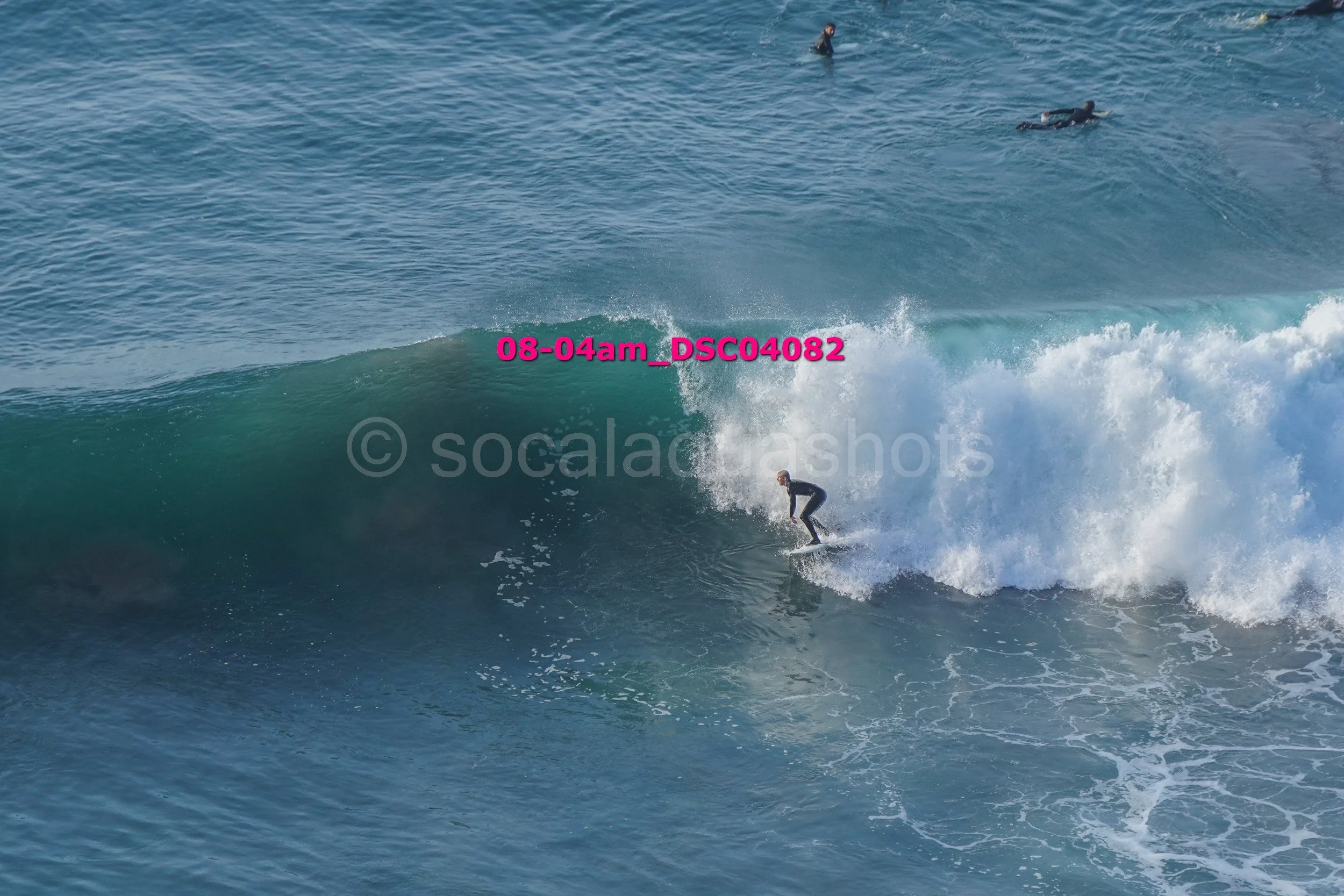 A surfer riding a large wave with two other surfers swimming in the water nearby.