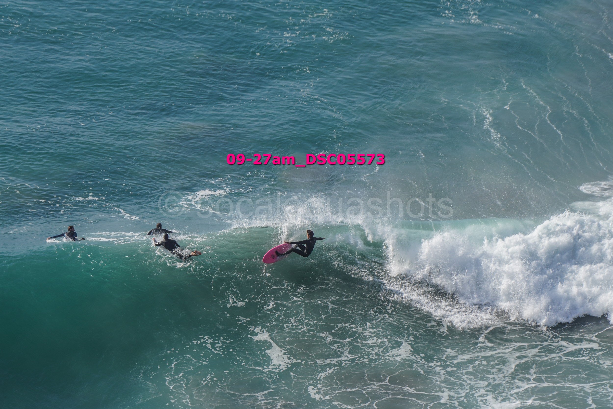 Surfer performing a jump on a pink surfboard on a large ocean wave, with two other surfers paddling nearby.