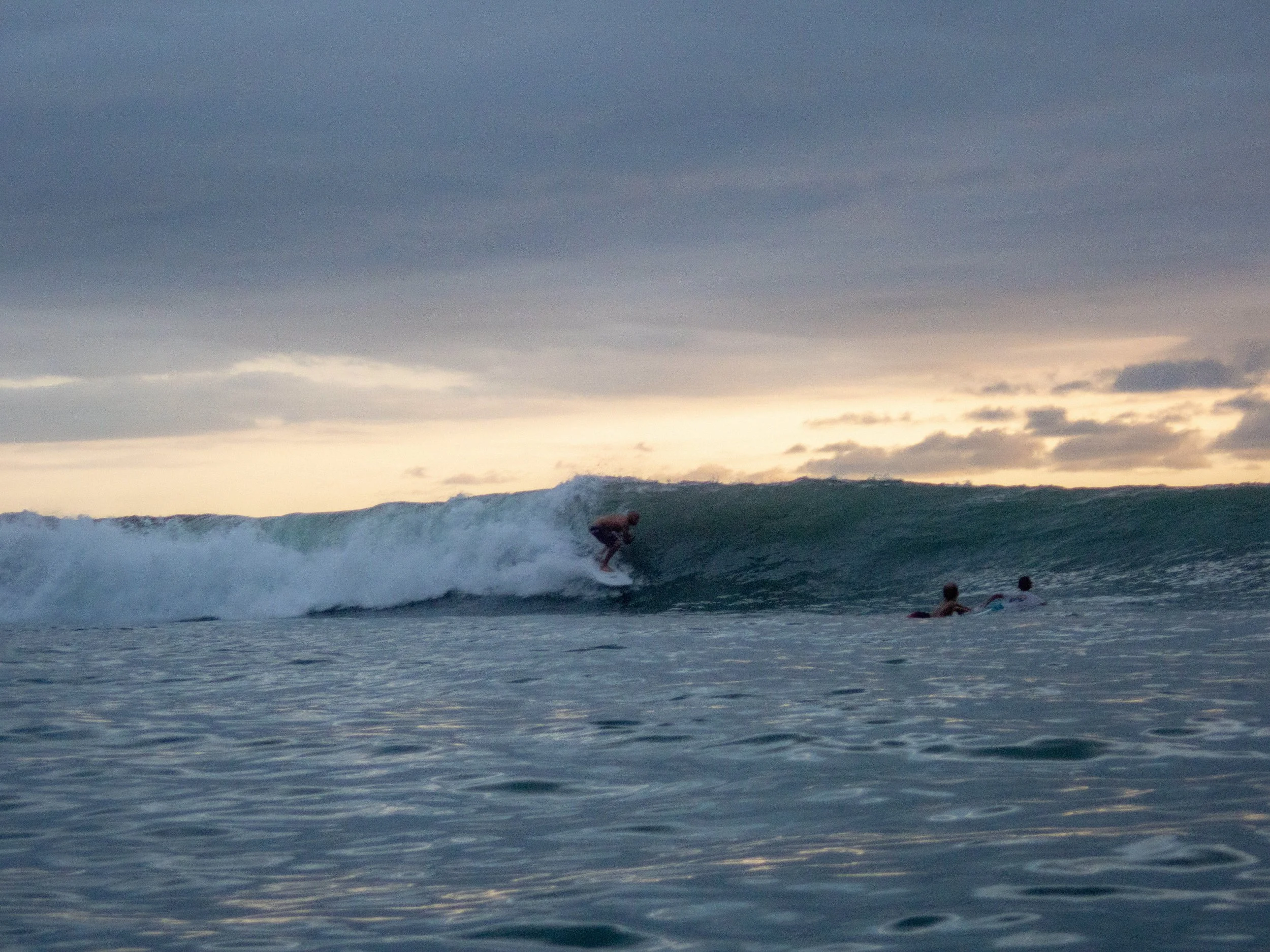Surfer riding a wave at sea with two people on surfboards nearby, during sunset, cloudy sky.
