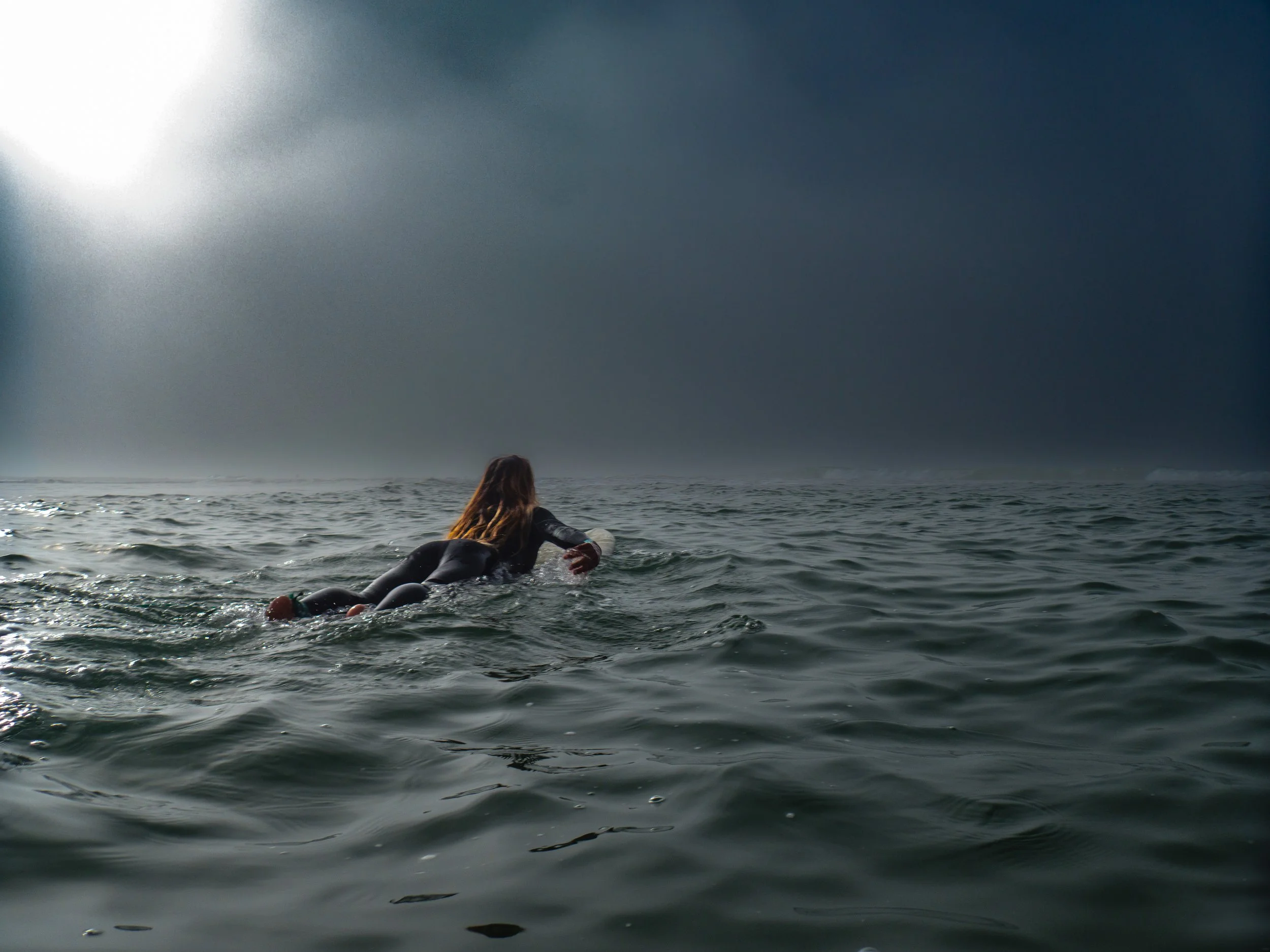 A person with long hair in a wetsuit paddling on a surfboard in open water under a cloudy sky.