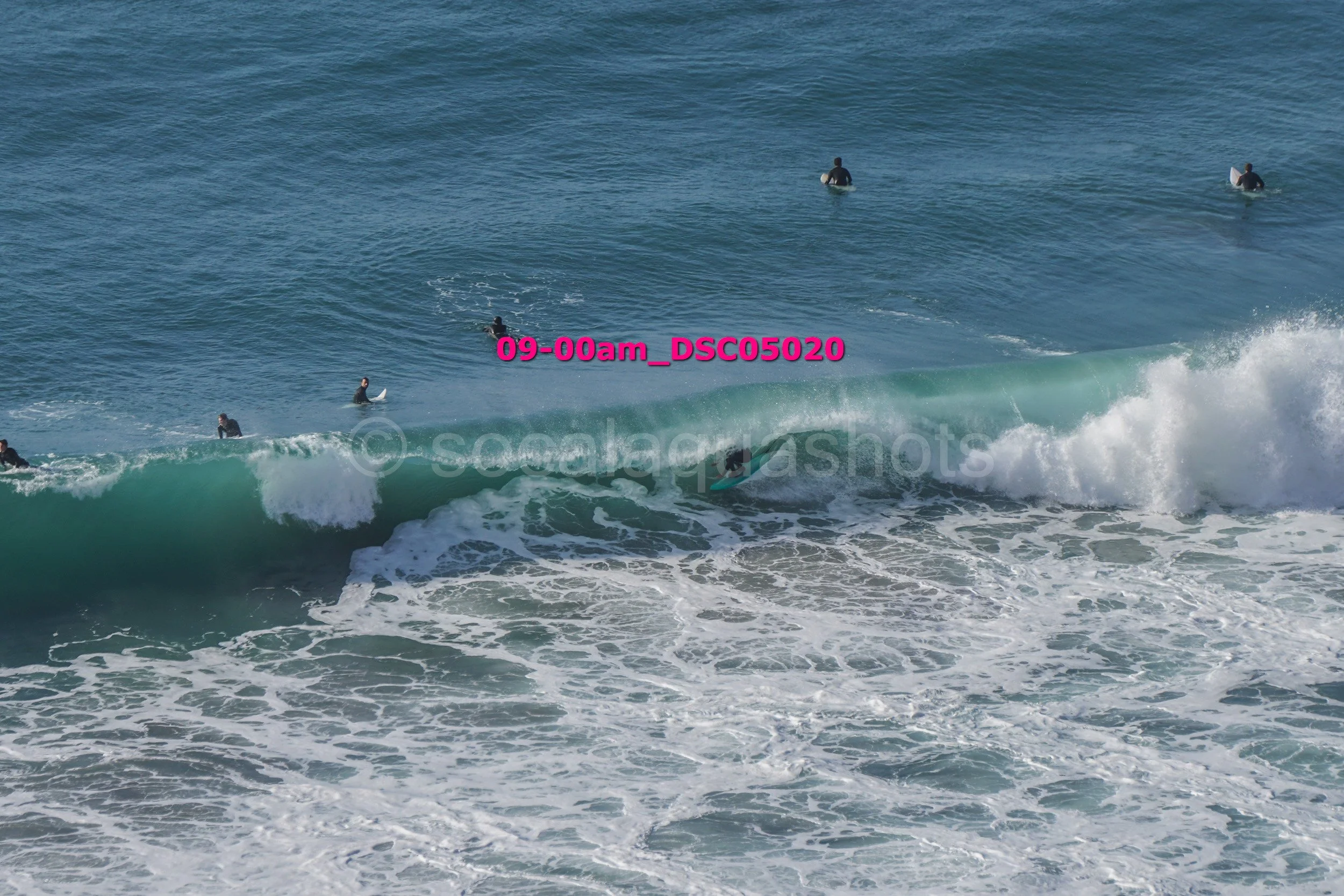 Surfers in the ocean, some riding a wave while others wait in the water, under a clear blue sky.