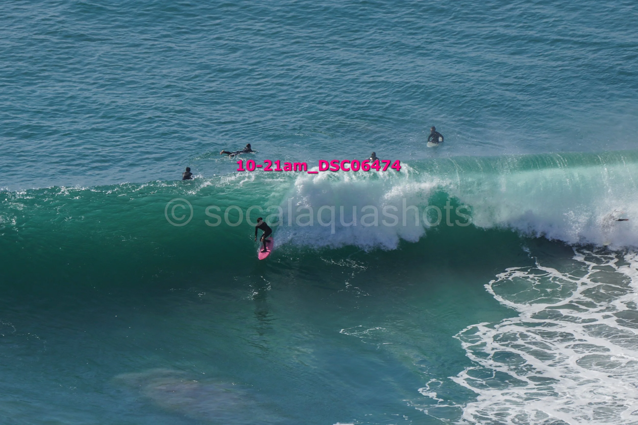 Surfer riding a large wave with five other surfers in the water nearby.