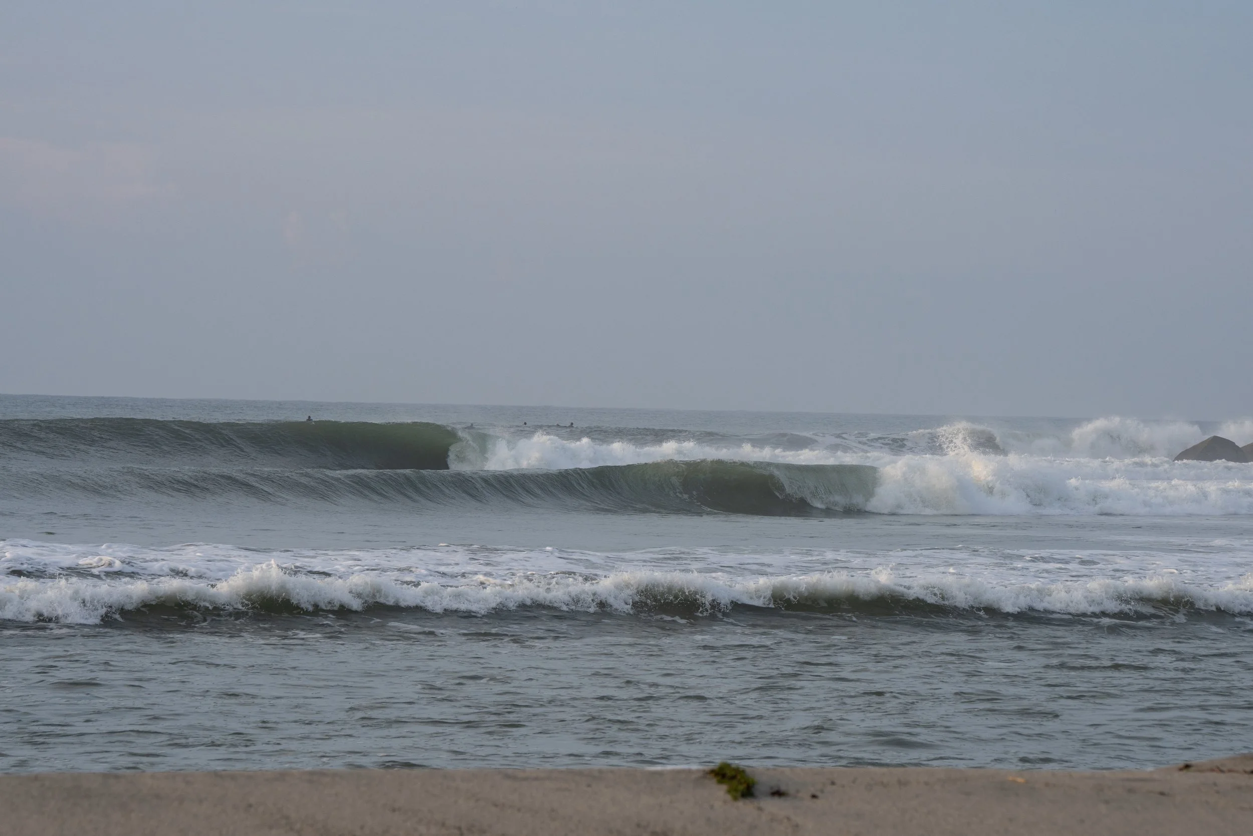 Sunny beach with gentle waves, a sandy shore, and a few people surfing in the background under a clear sky.
