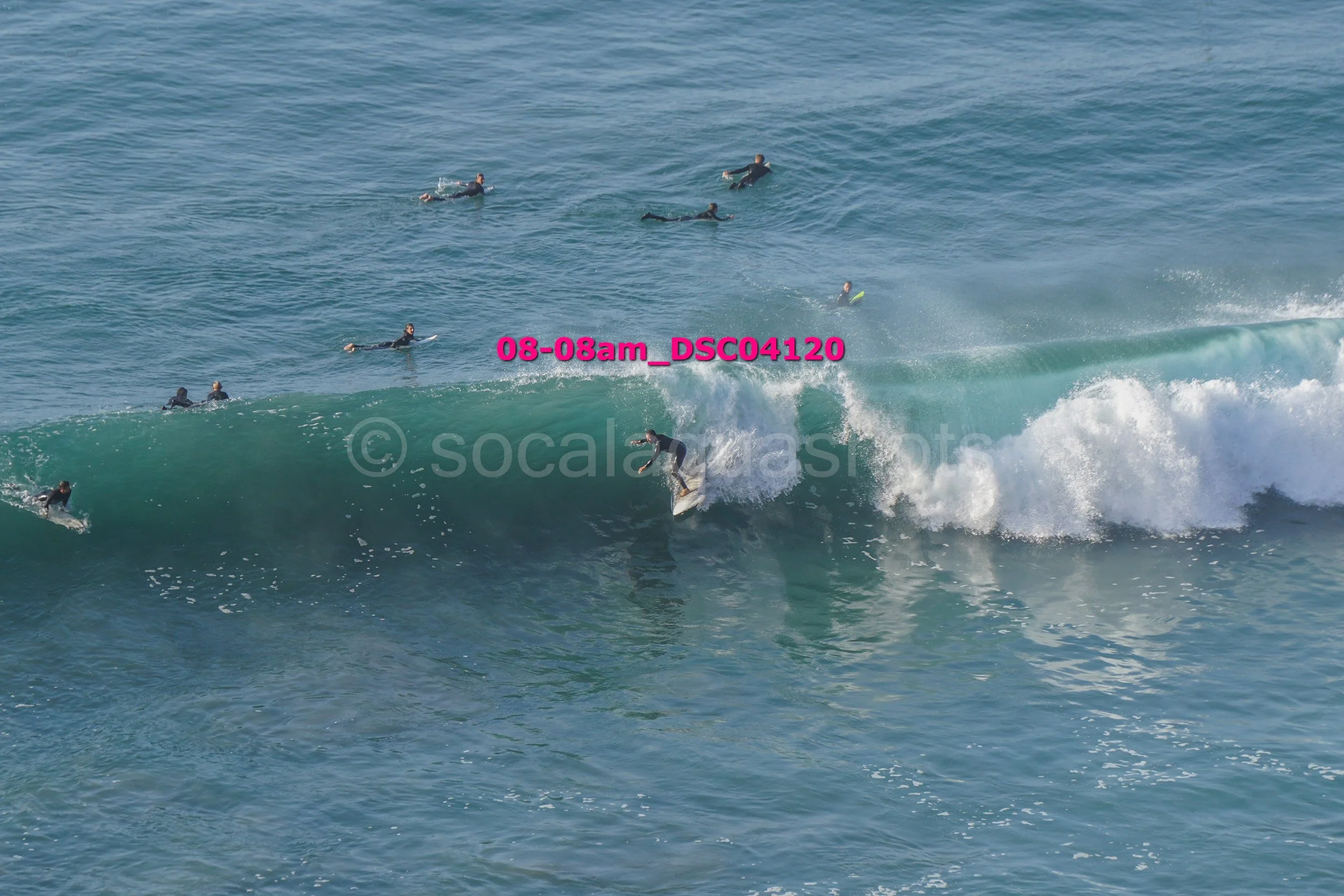 Surfer riding a large wave with several people swimming and floating in the water nearby.