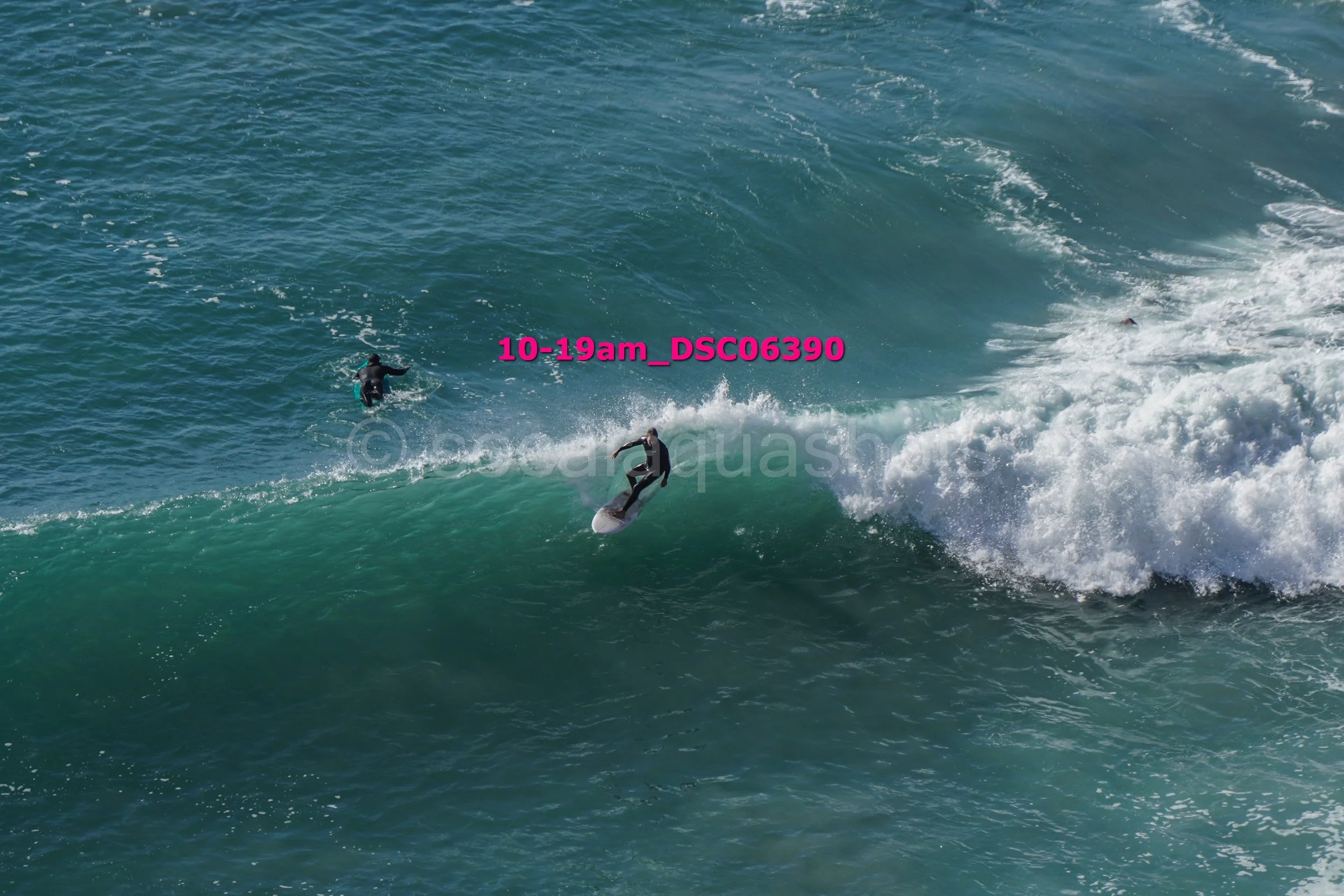 Surfer riding a wave in the ocean with another person paddling nearby.
