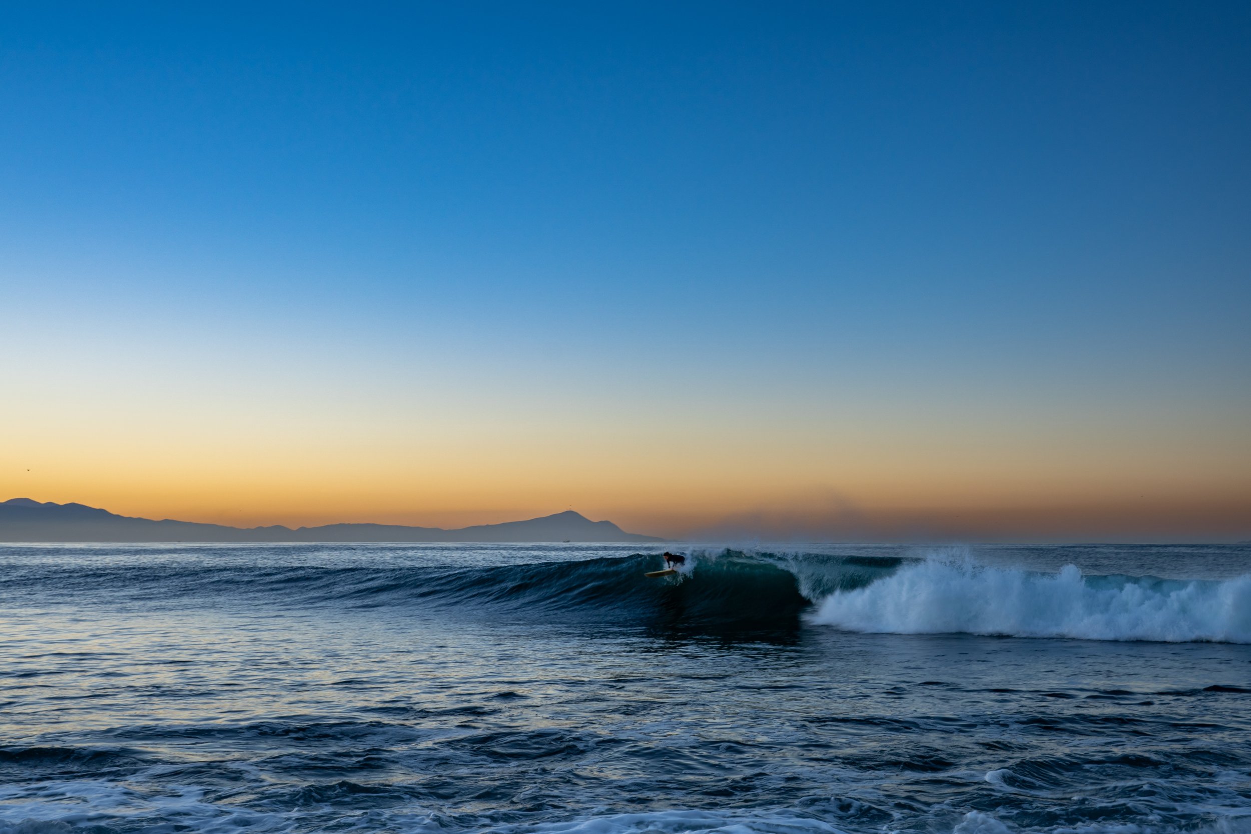 A surfer riding a wave in the ocean during sunset with a mountain range in the background.