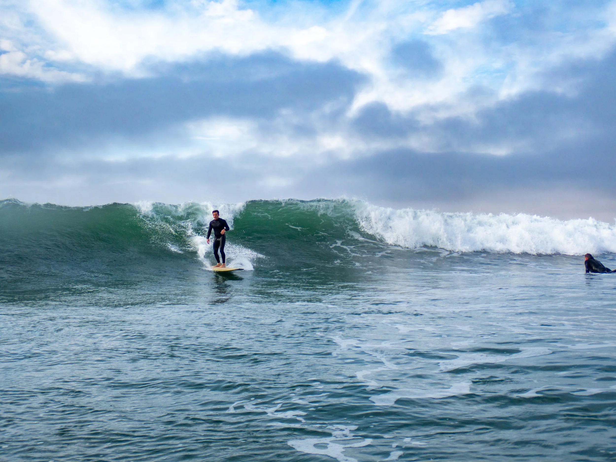 A man surfing a wave in the ocean with another surfer lying on their board nearby under a cloudy sky.