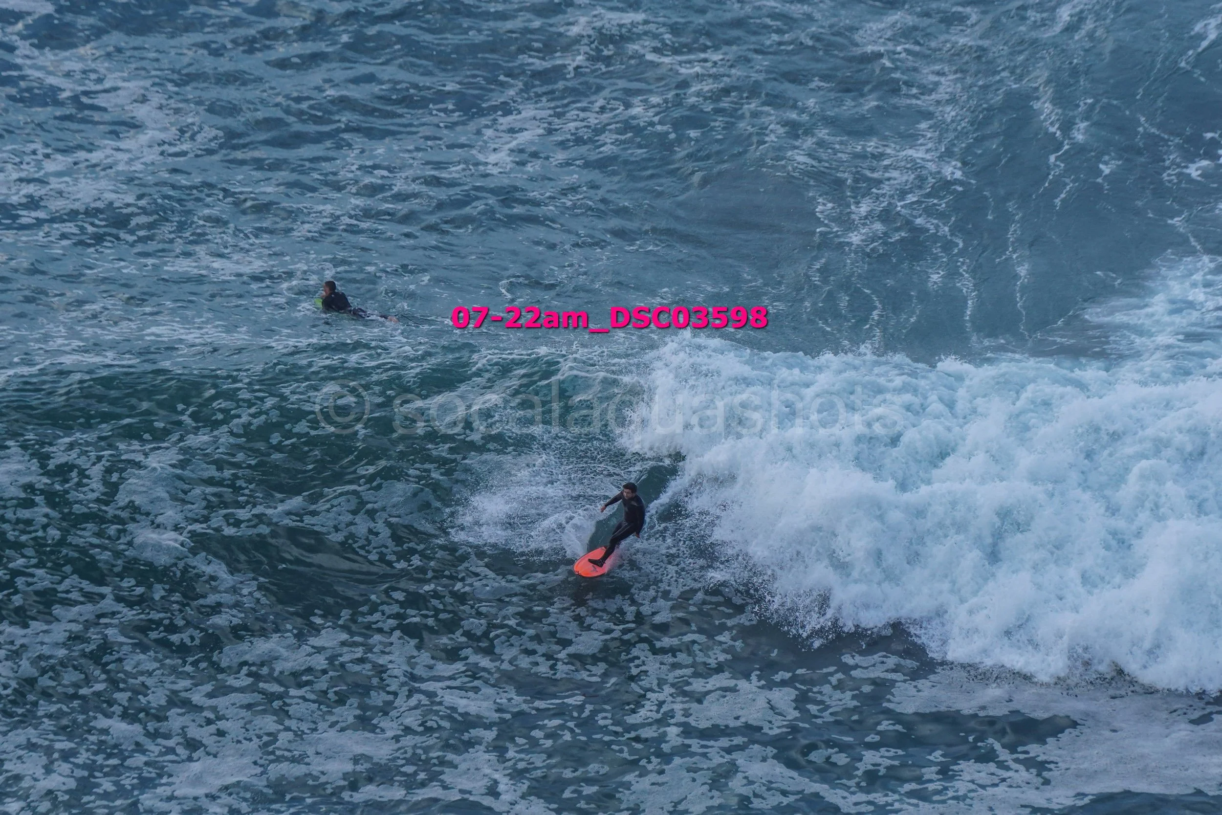Two surfers in wetsuits riding and swimming in the ocean waves, with one actively surfing and the other paddling in the water.