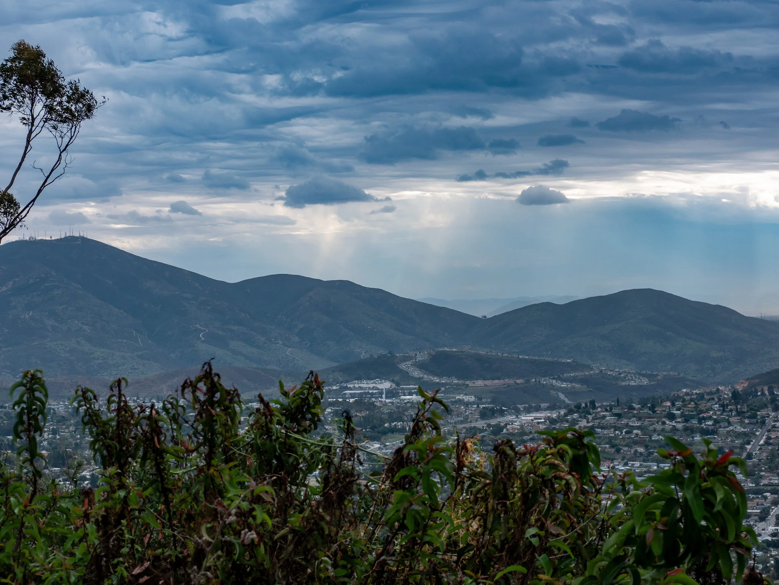 Landscape view of distant mountain range under cloudy sky, with urban area and greenery in the foreground.