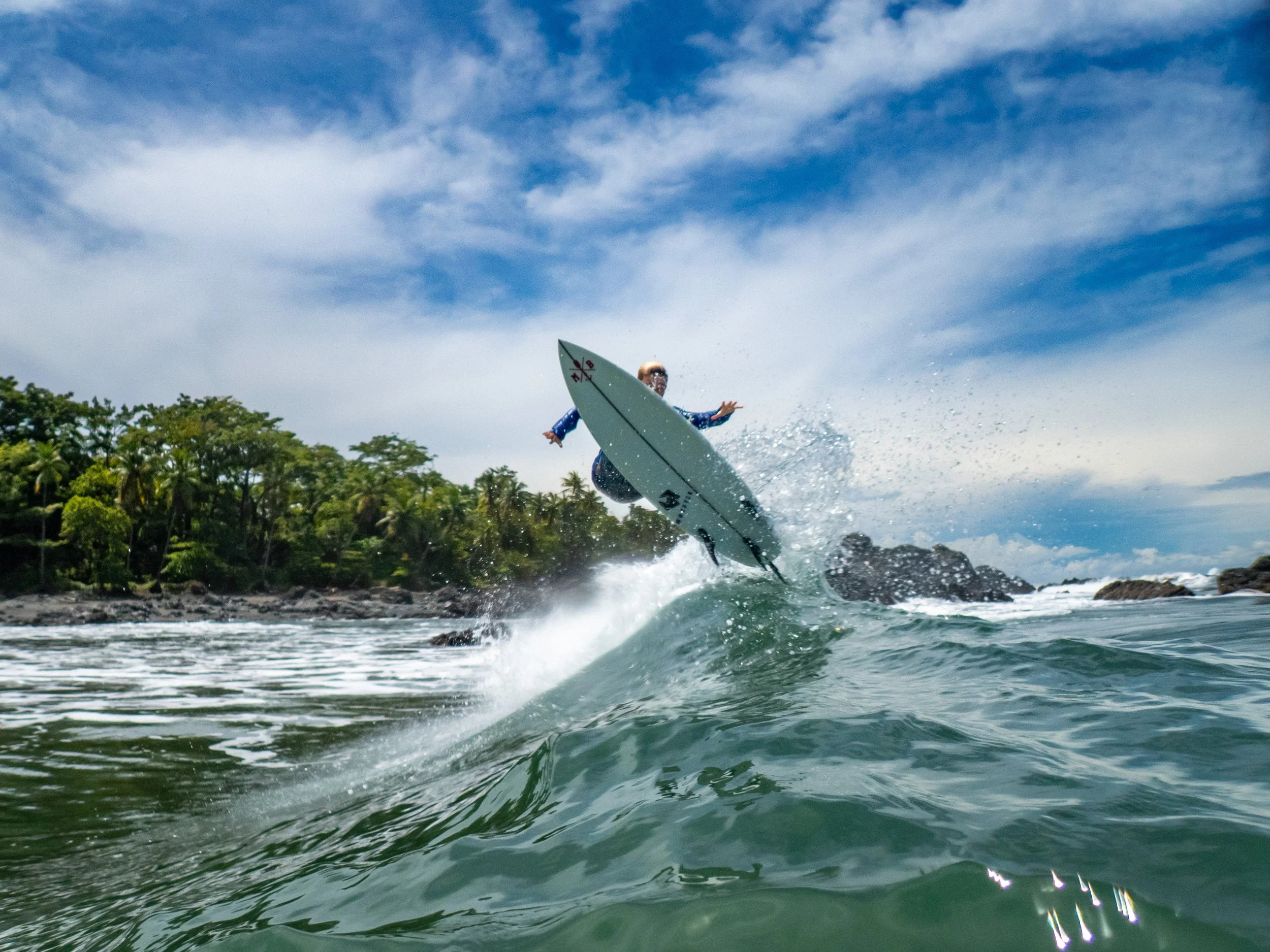 Surfer riding a wave near a tropical coastline with lush green trees and rocks under a partly cloudy sky.