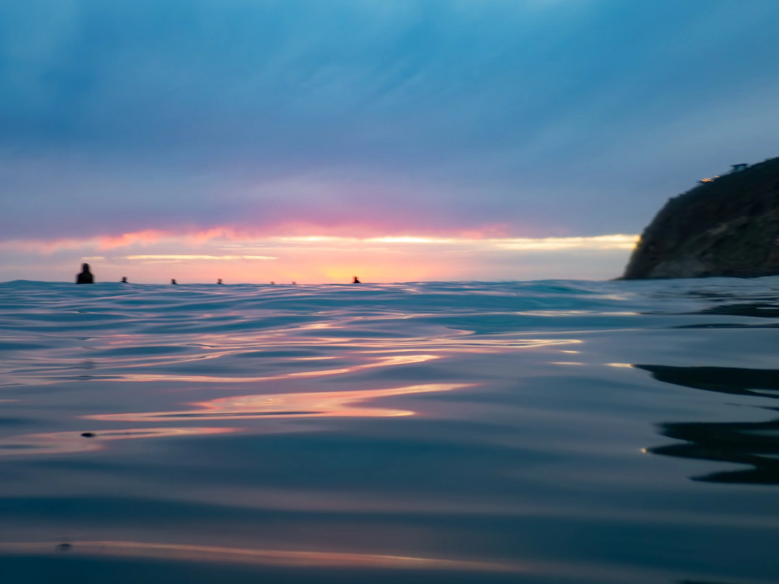 Calm ocean water at sunset with a distant silhouette of a cliff on the right and a few swimmers in the distance, under a colorful sky with clouds.