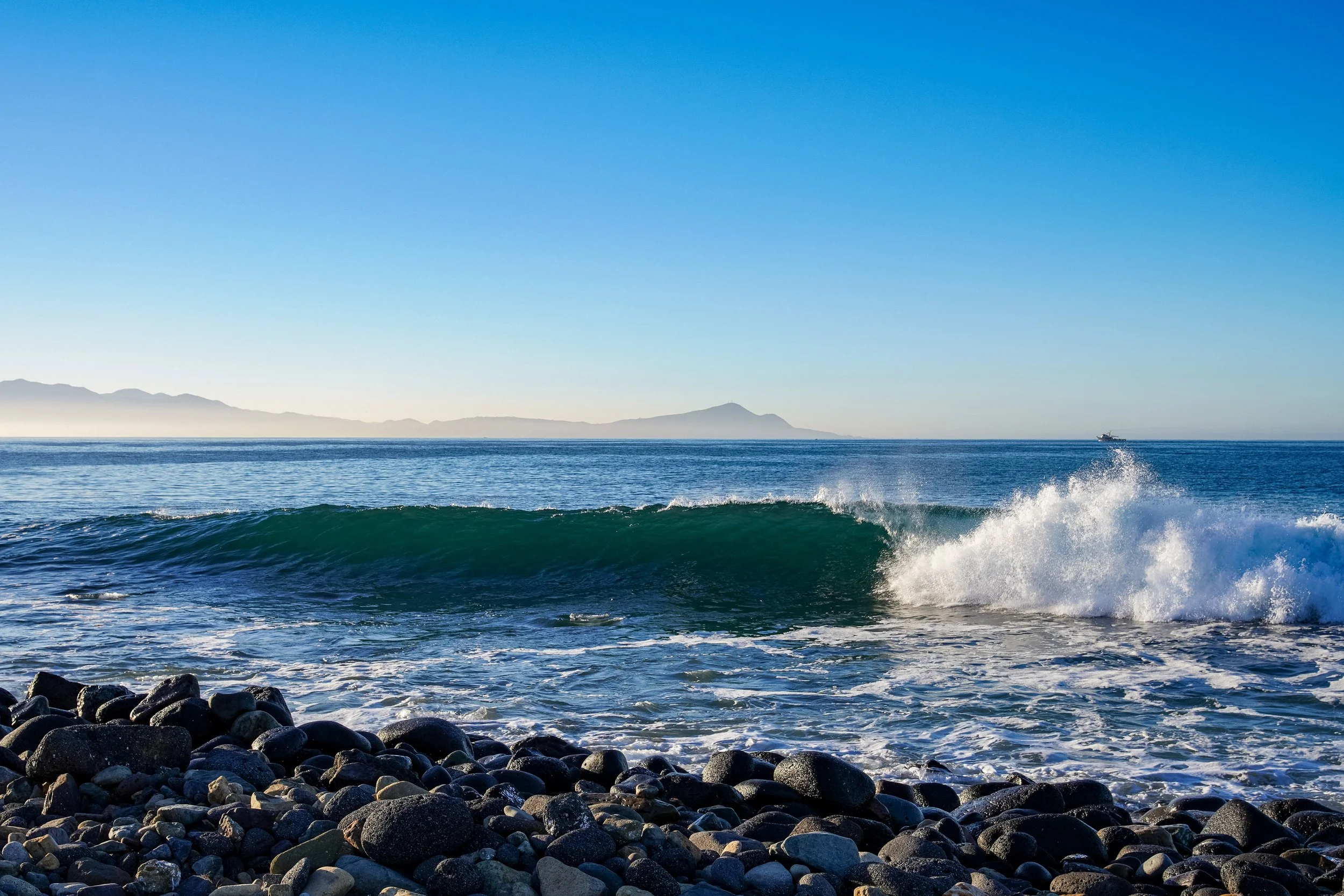 Ocean waves crashing onto a rocky shoreline with mountains in the background and a boat on the water under a clear blue sky.