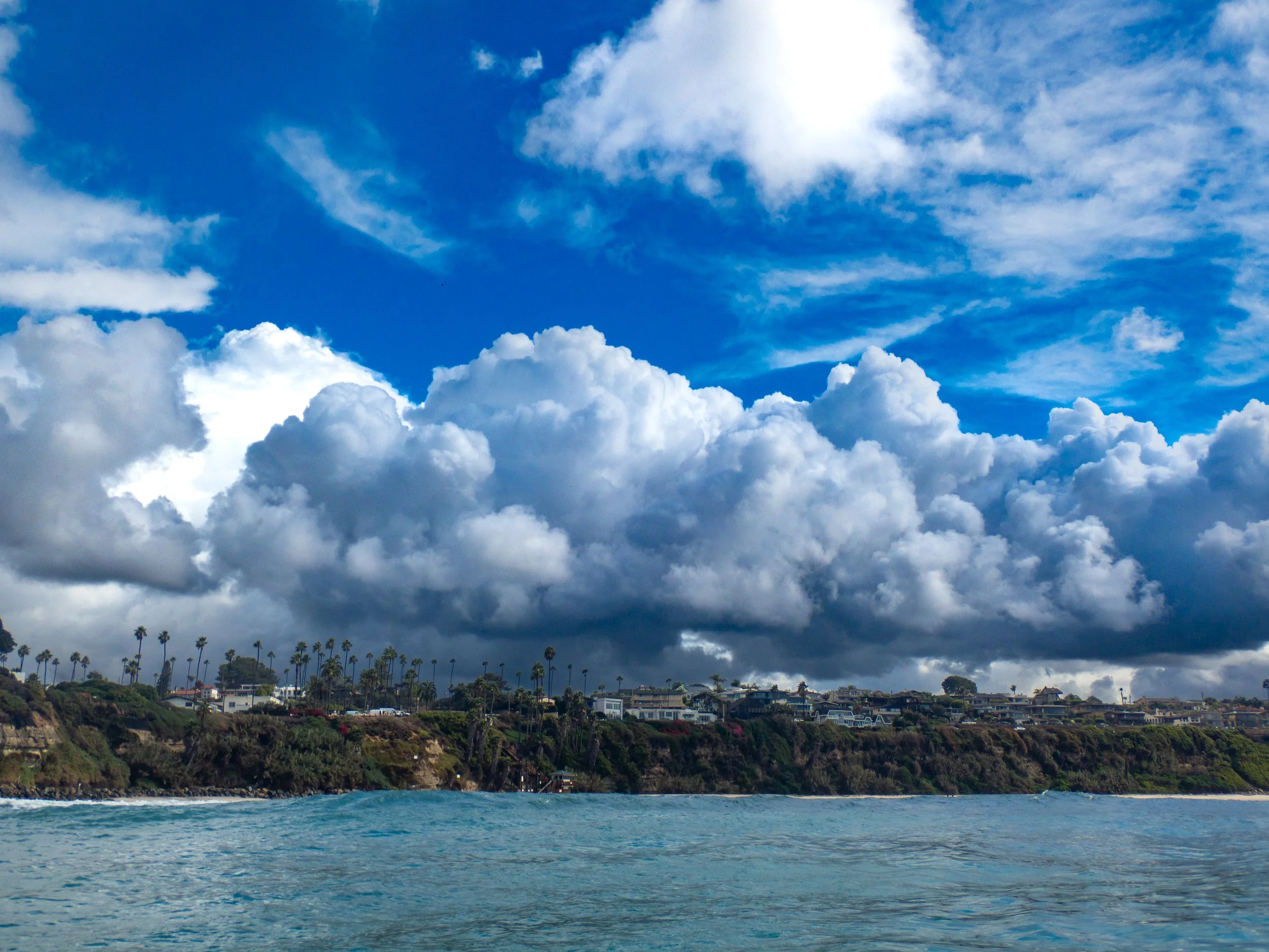 Ocean with blue water in foreground, topped with a coastal hillside covered with trees and houses, and a large sky filled with fluffy white clouds and some darker clouds.