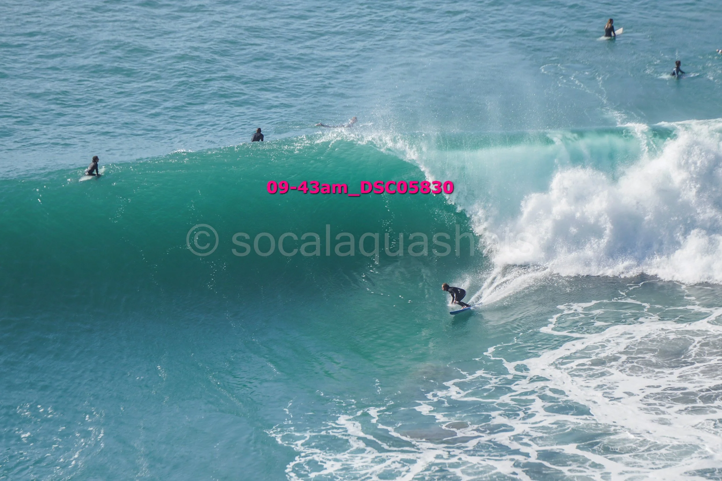 A surfer riding a large wave at the beach with multiple people surfing and swimming in the background.