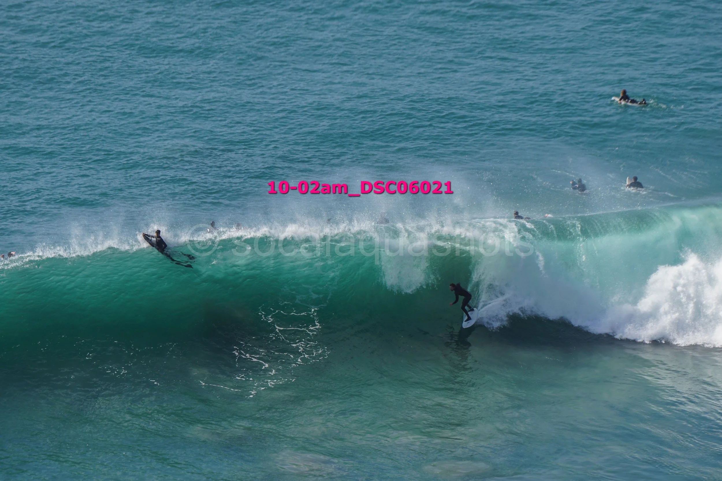 Surfer riding a large green wave with multiple surfers in the water in the background.