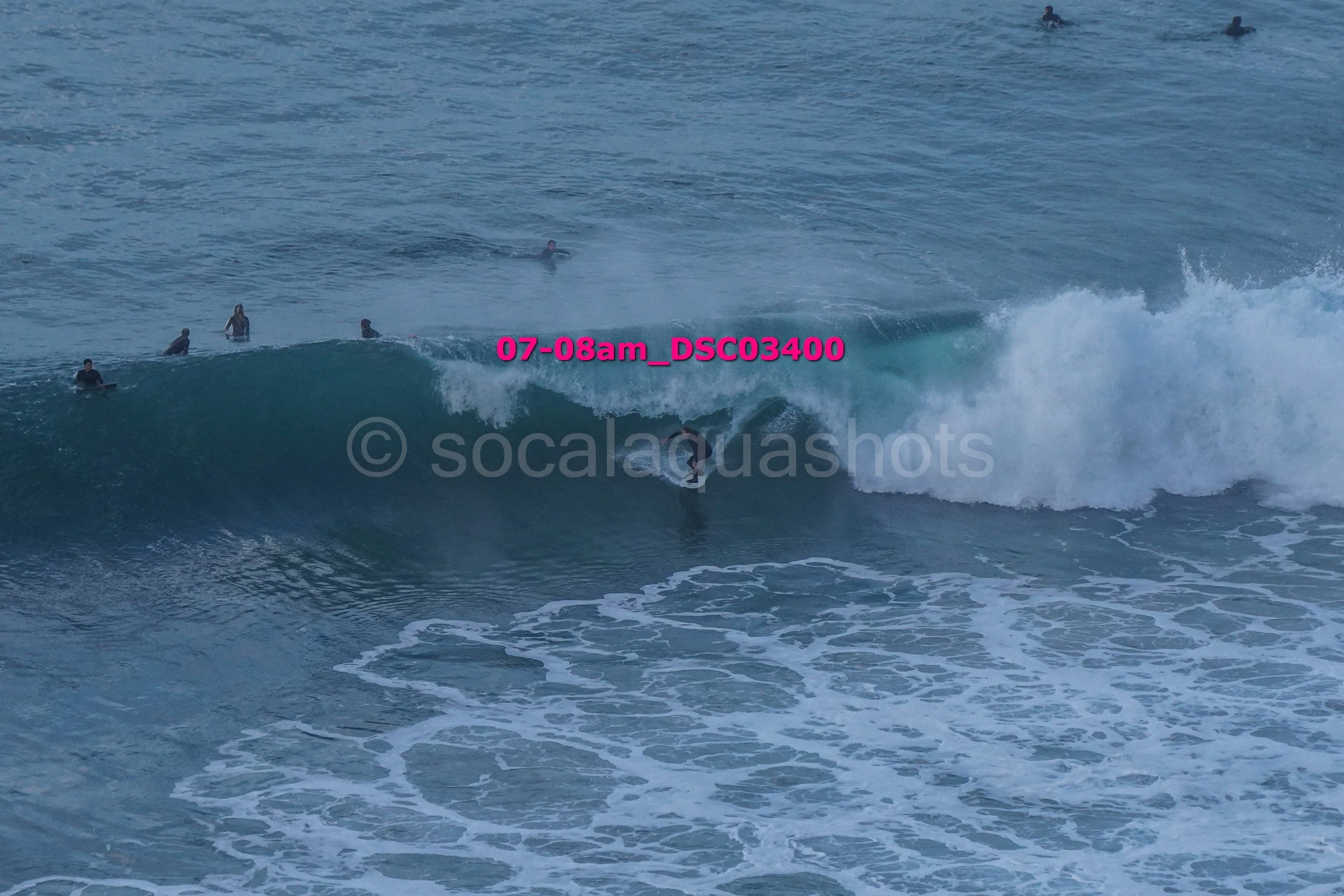 Surfer riding a wave with several surfers watching from the water in the background at the beach.