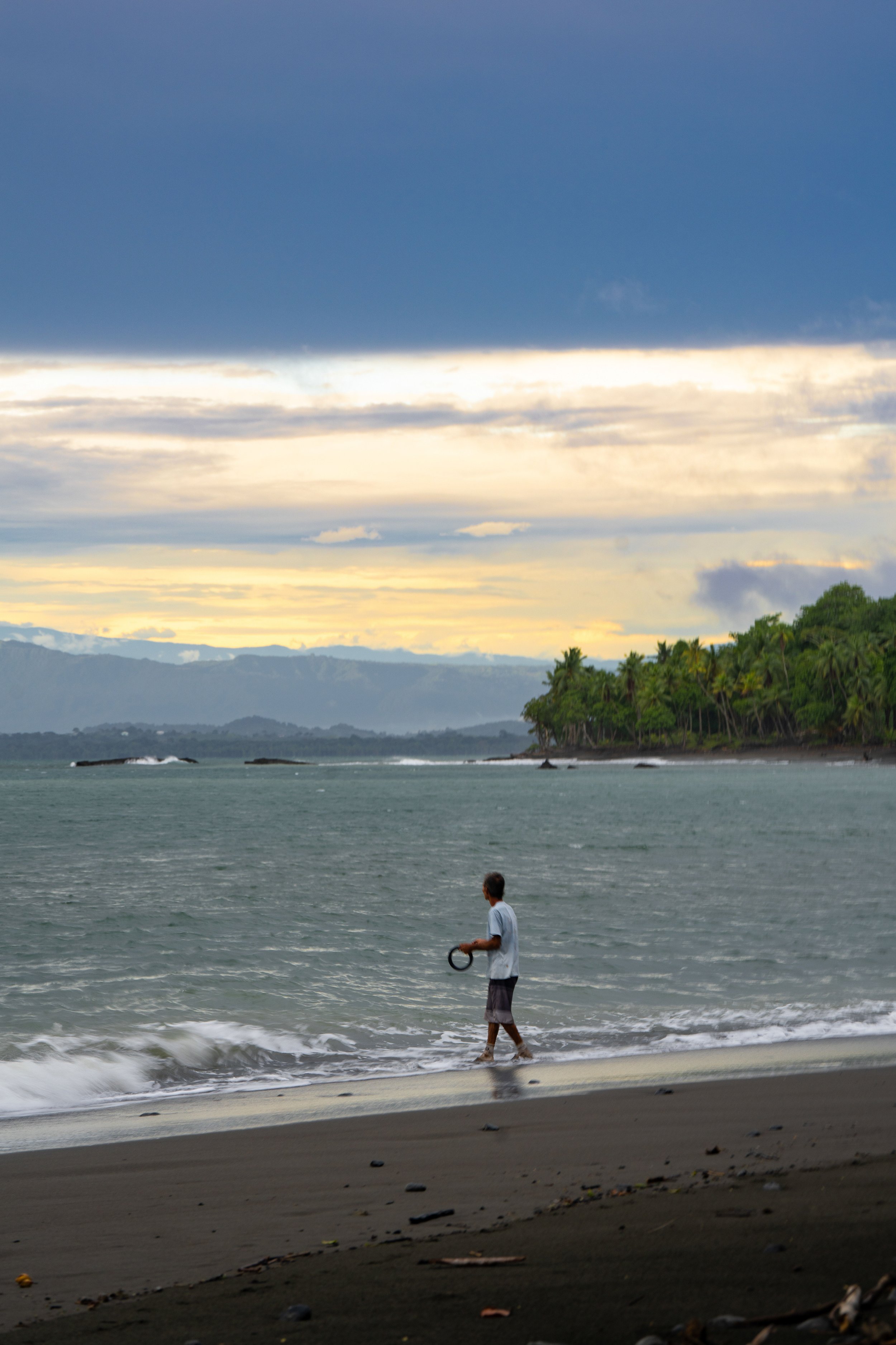 A person walking on the beach near the water with trees and hills in the background under a cloudy sky.