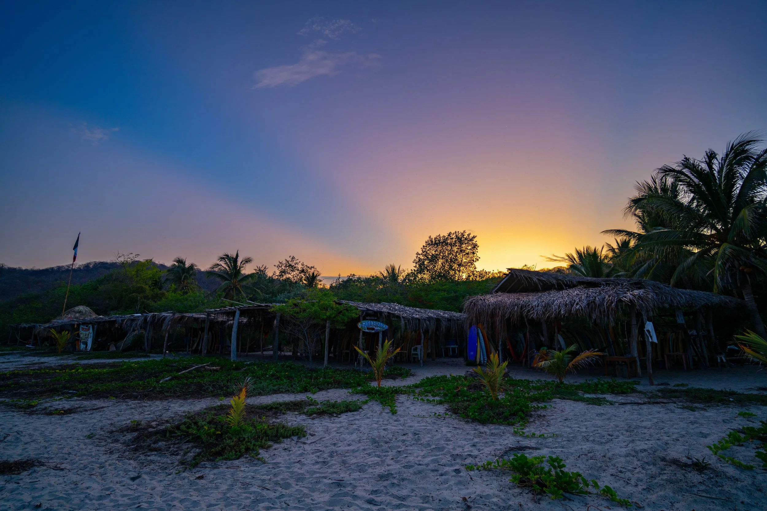 A tropical beach scene at sunset with a clear sky, palm trees, and thatched huts.
