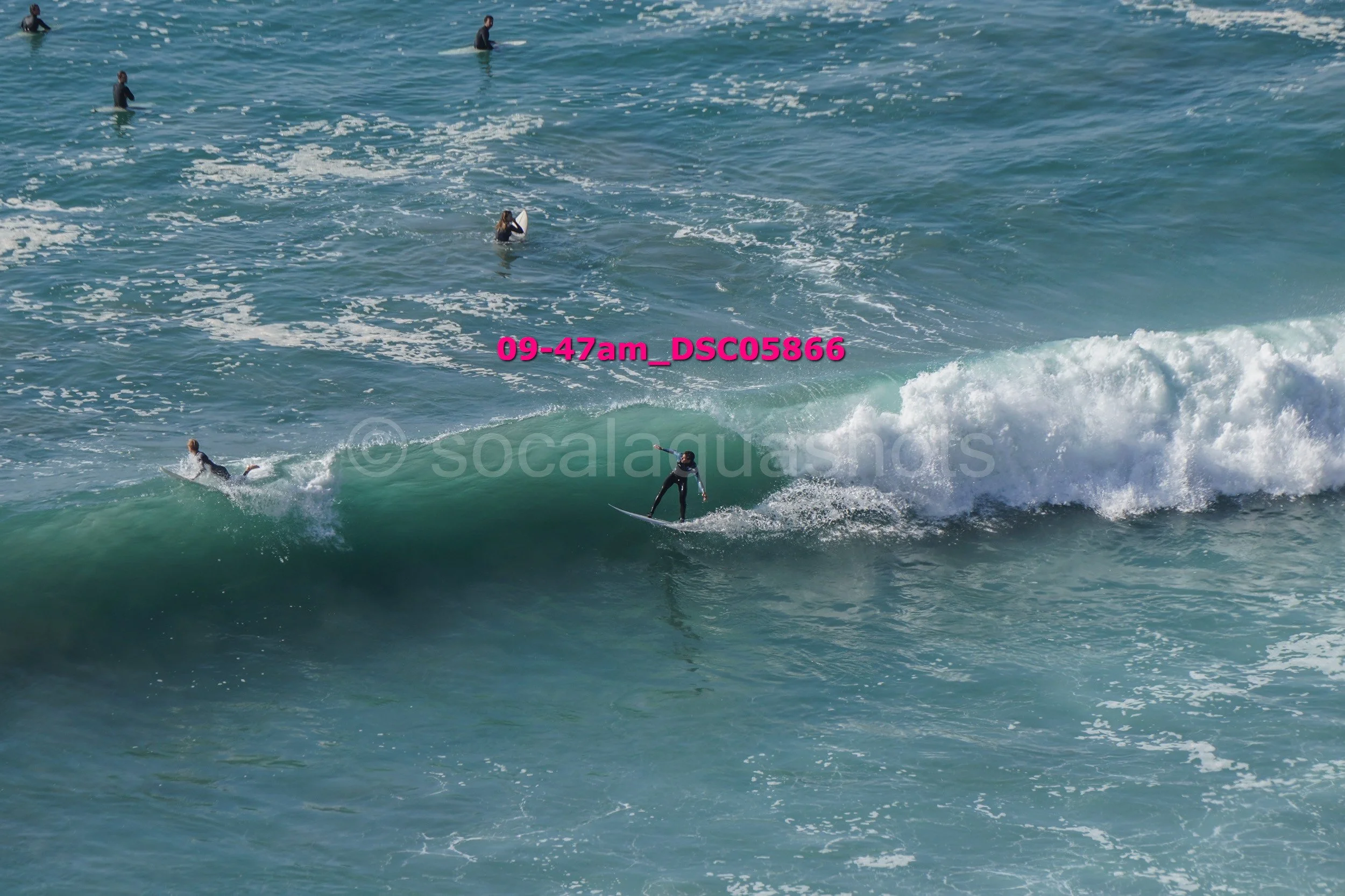 Surfers in the ocean, some riding waves while others wait or paddle, during daylight hours.