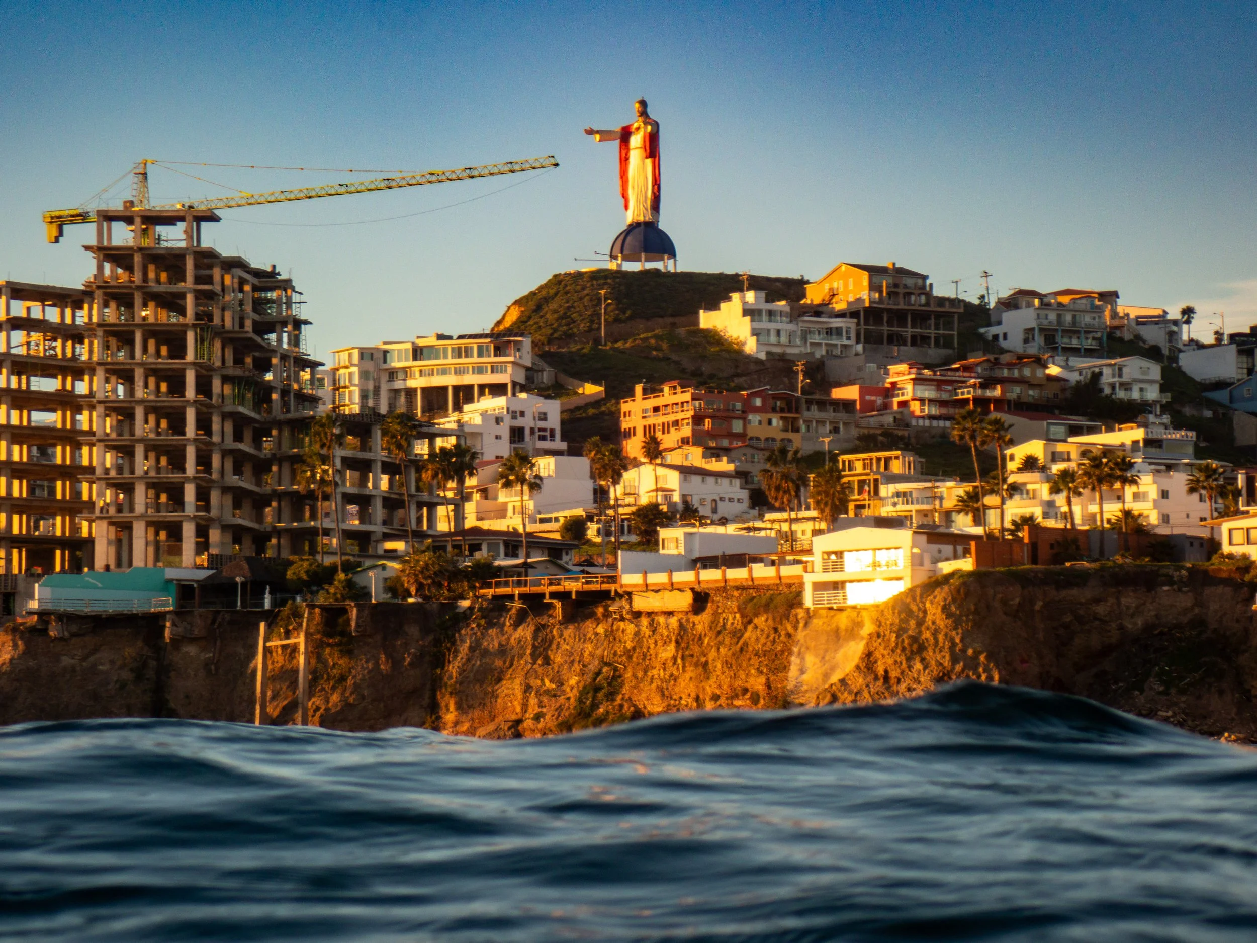 View of the city of San Diego at sunset, with the Christ the Redeemer statue on a hill in the background, and the ocean in the foreground.