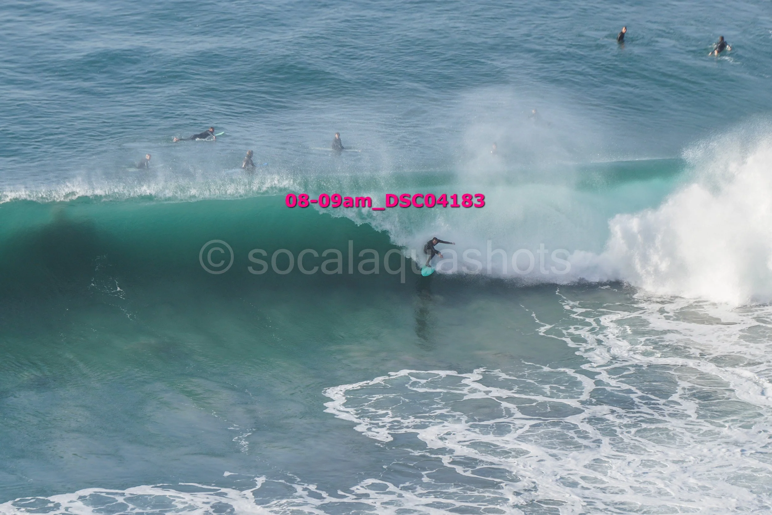 A person surfing on a large ocean wave with several other surfers in the background.