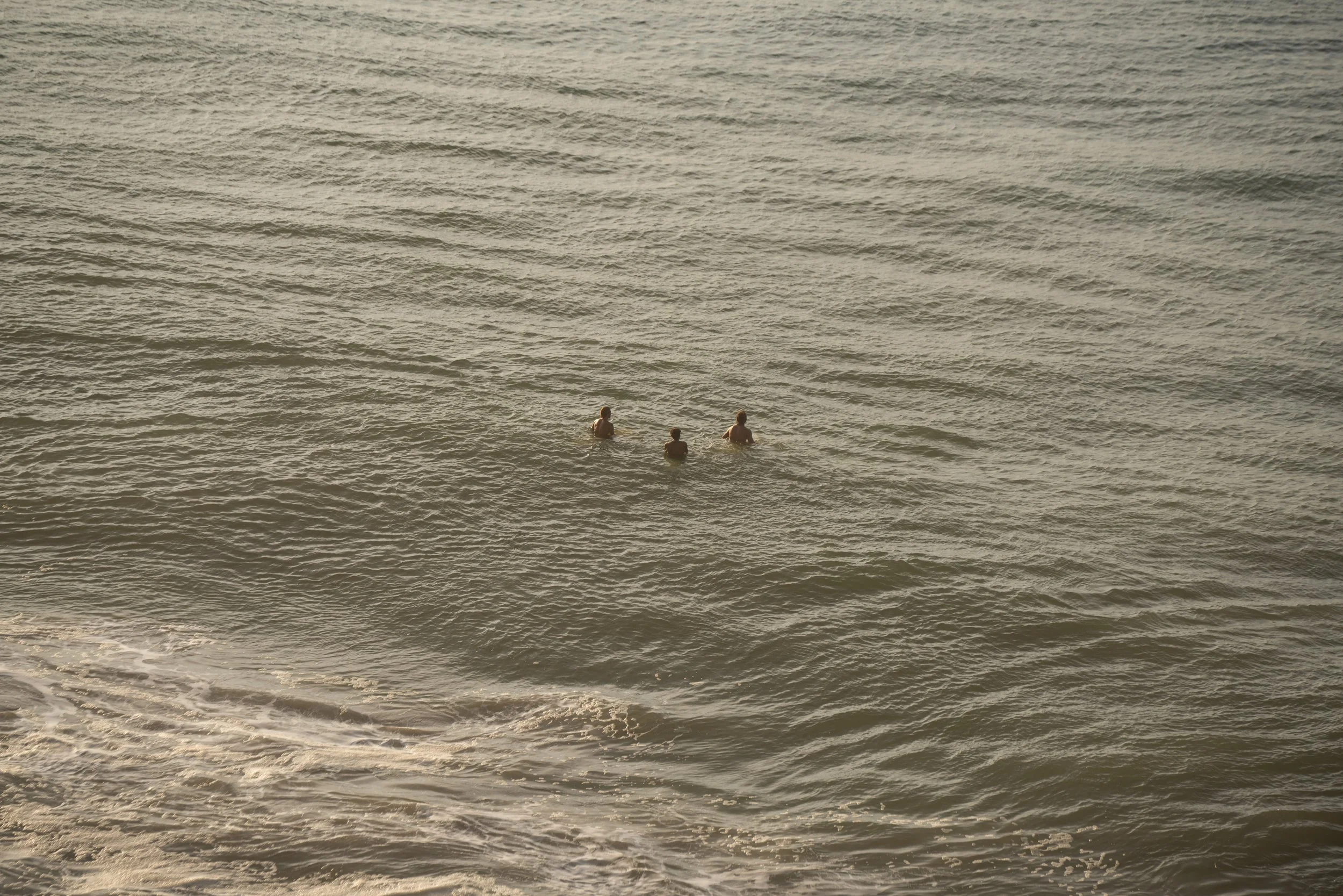 Three people swimming in the ocean near the shore during sunset or sunrise with calm waves.