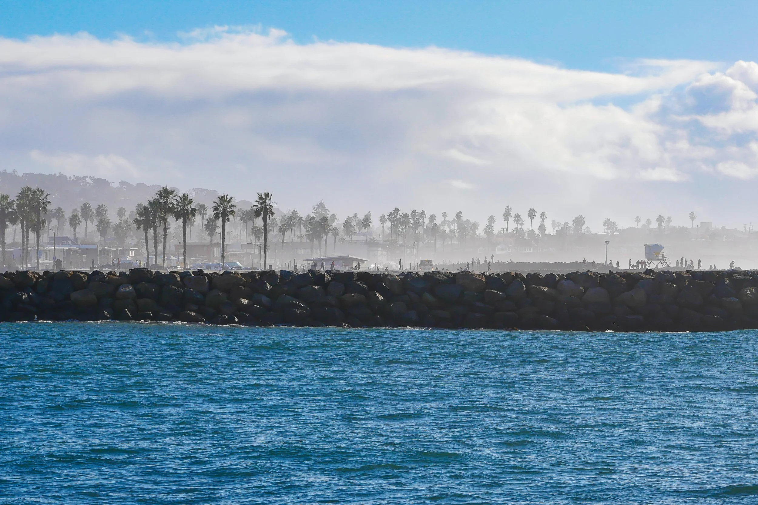 Ocean waves in foreground, rocky breakwater with silhouetted people and palm trees in background, cloudy sky.