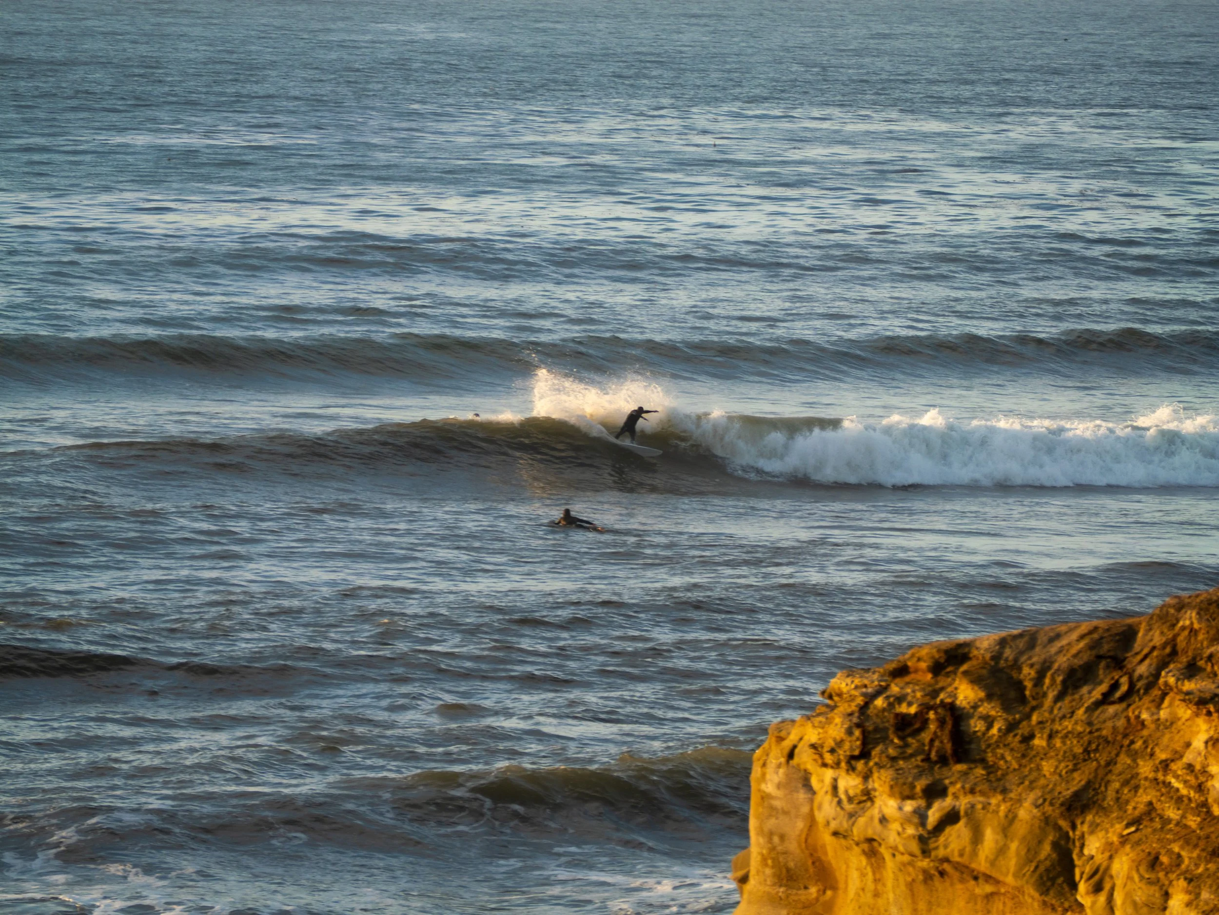 A person surfing on a wave in the ocean with another person swimming nearby and a rocky shoreline in the foreground.