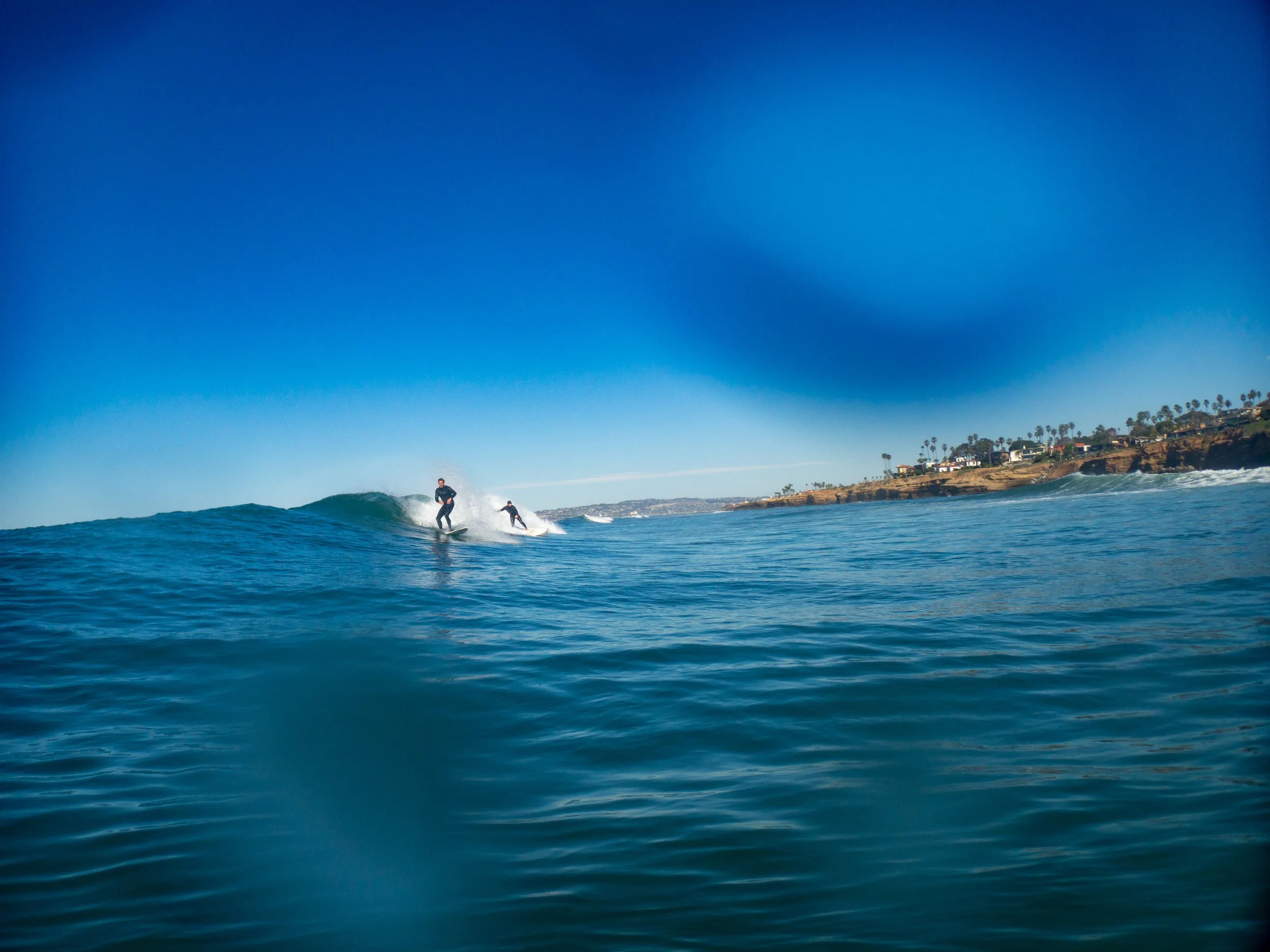 Two surfers riding a wave in the ocean near a coastline with houses and palm trees under a clear blue sky.