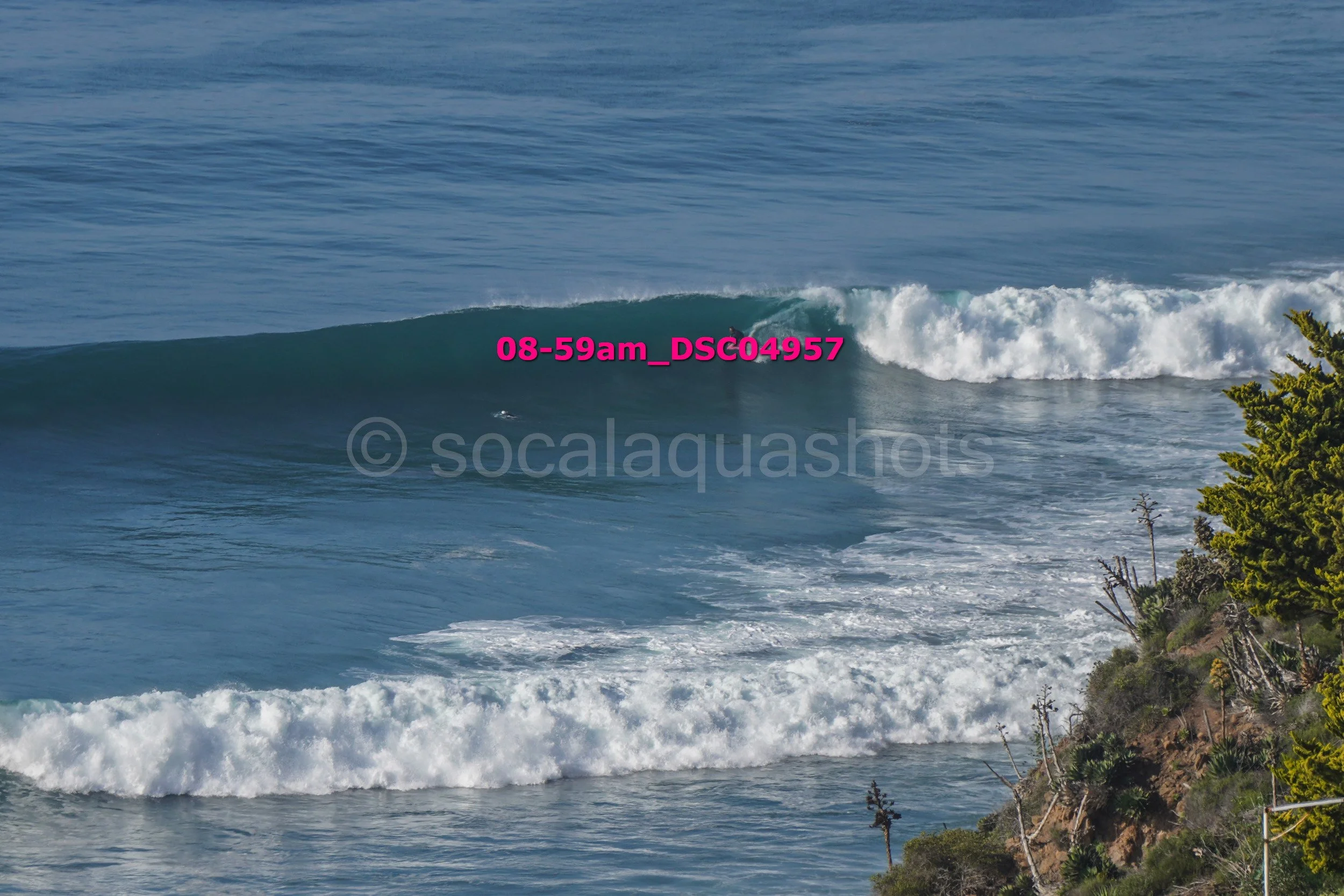 Ocean waves breaking near a rocky shoreline with green trees on the right side.