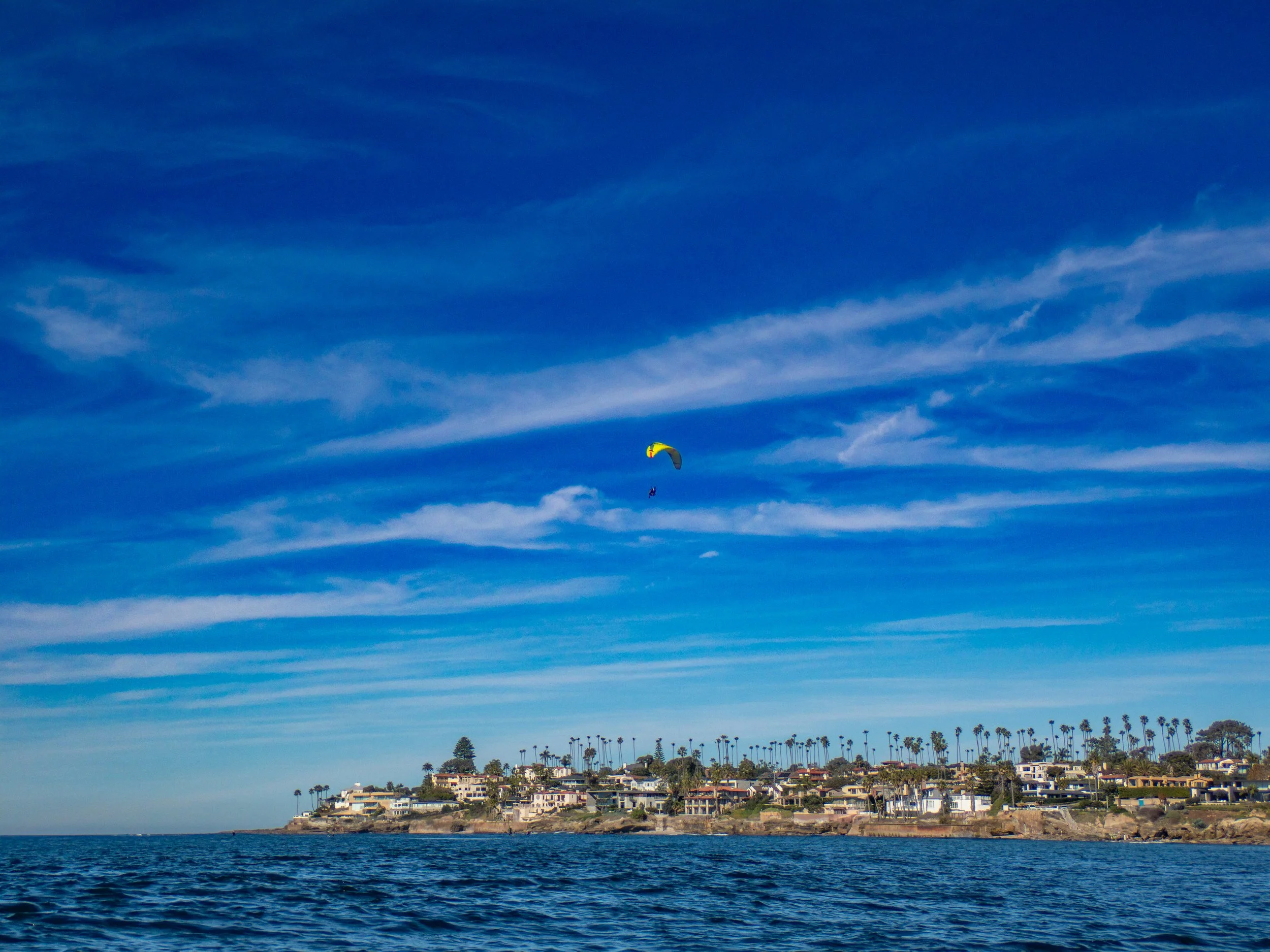 A person paragliding over a coastal neighborhood with houses and palm trees, with a bright blue sky and wispy clouds above.