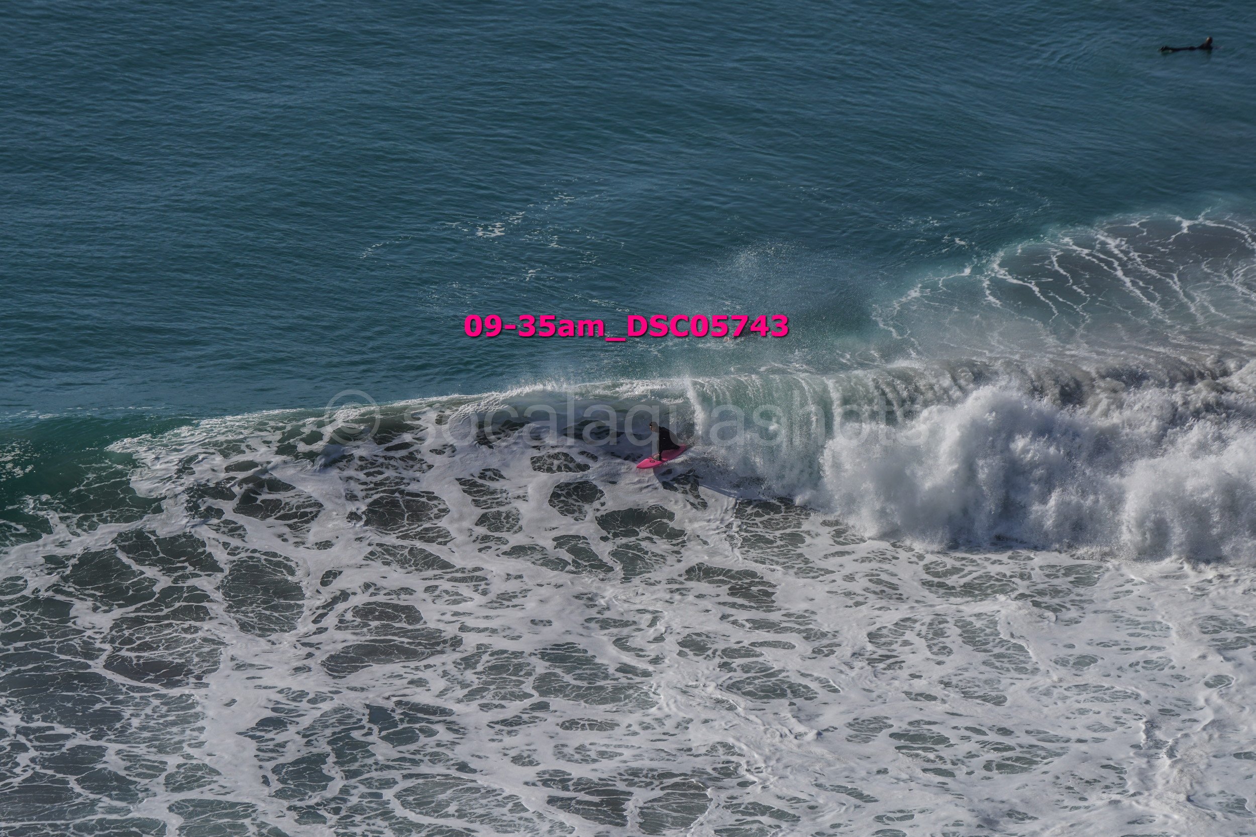 A person surfing on a pink surfboard in the ocean with waves breaking around them, and a small boat in the background.
