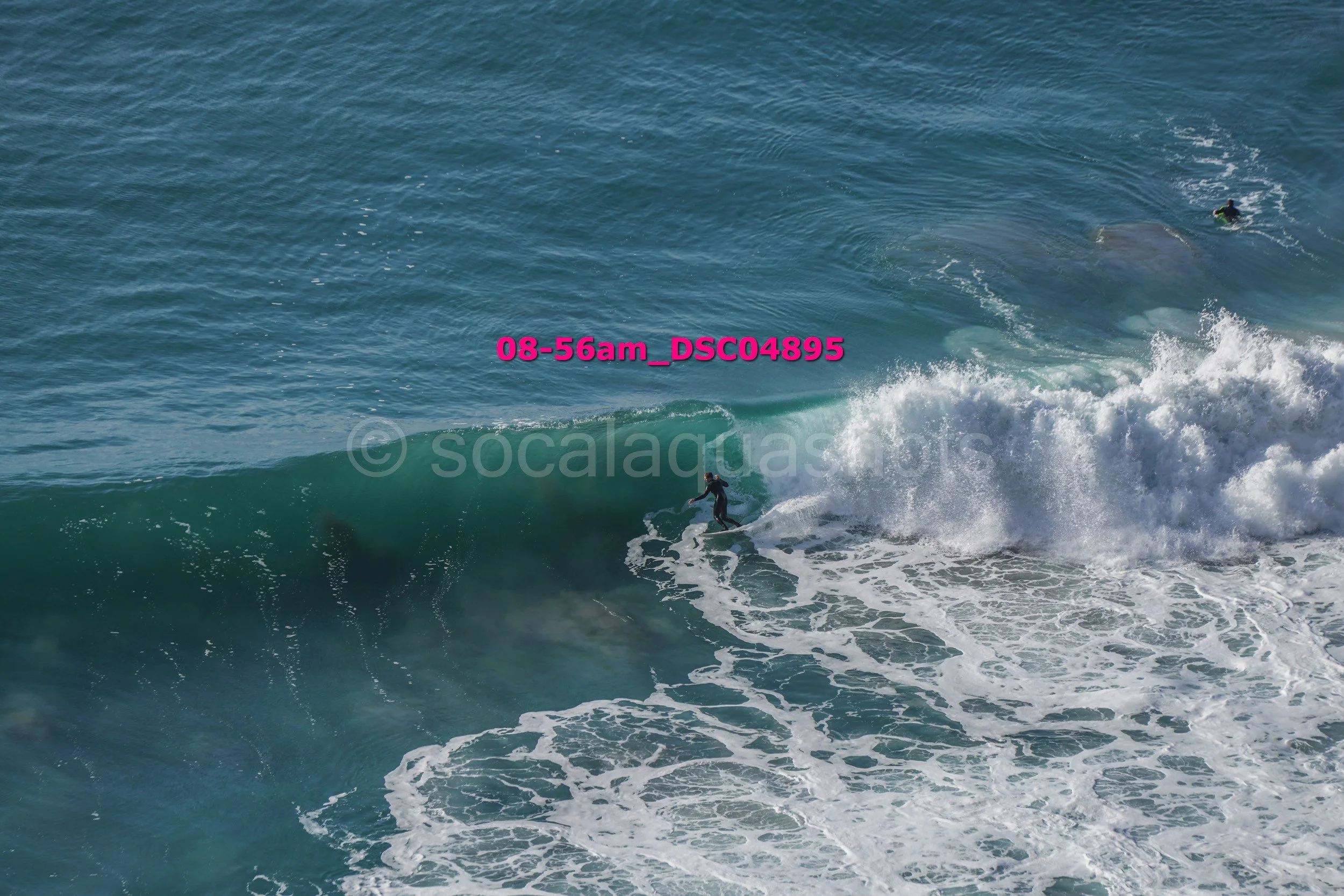 A person surfing on a wave in the ocean, with another surfer visible in the distance.
