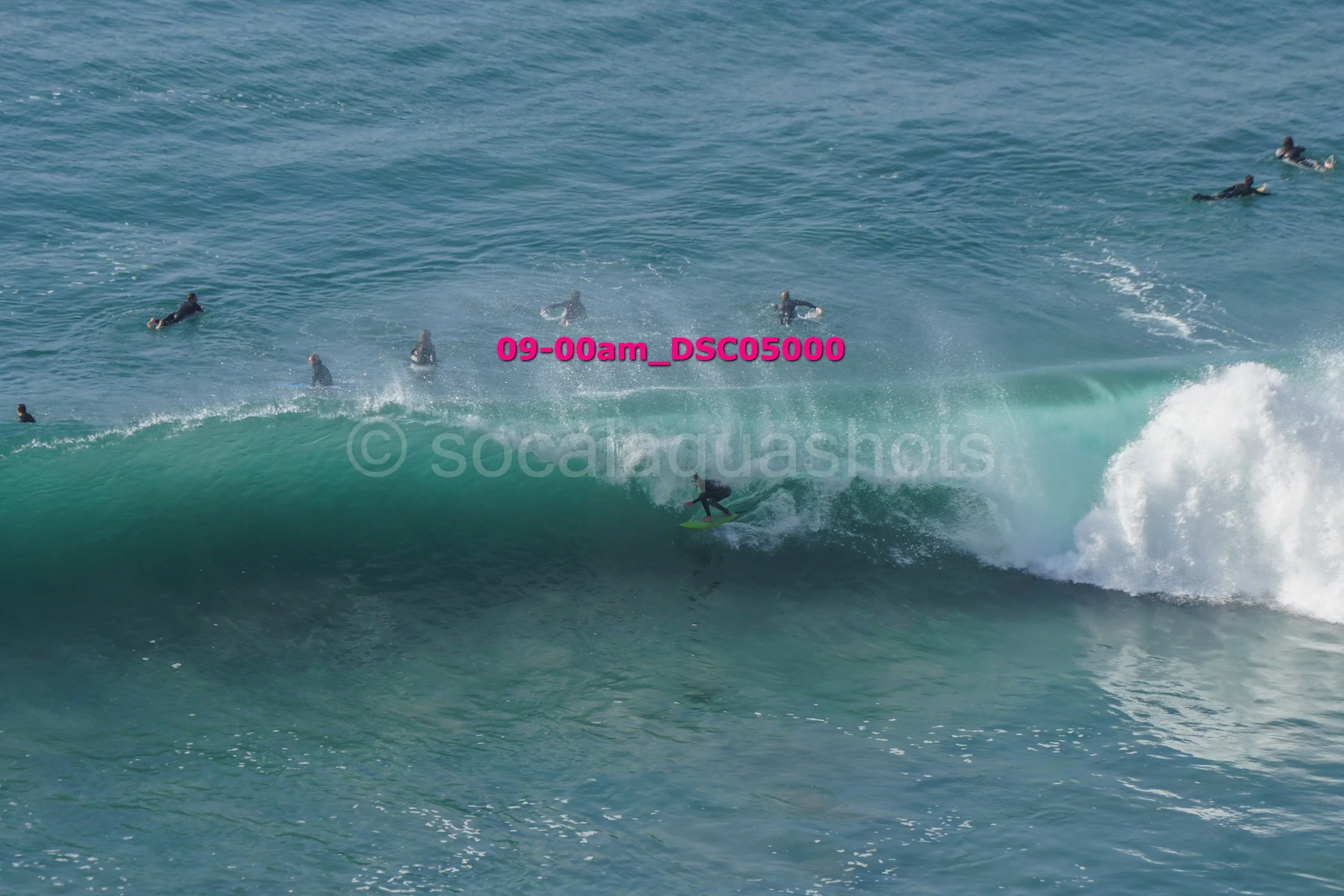 Surfer riding a large wave with multiple people swimming and surfing in the background.