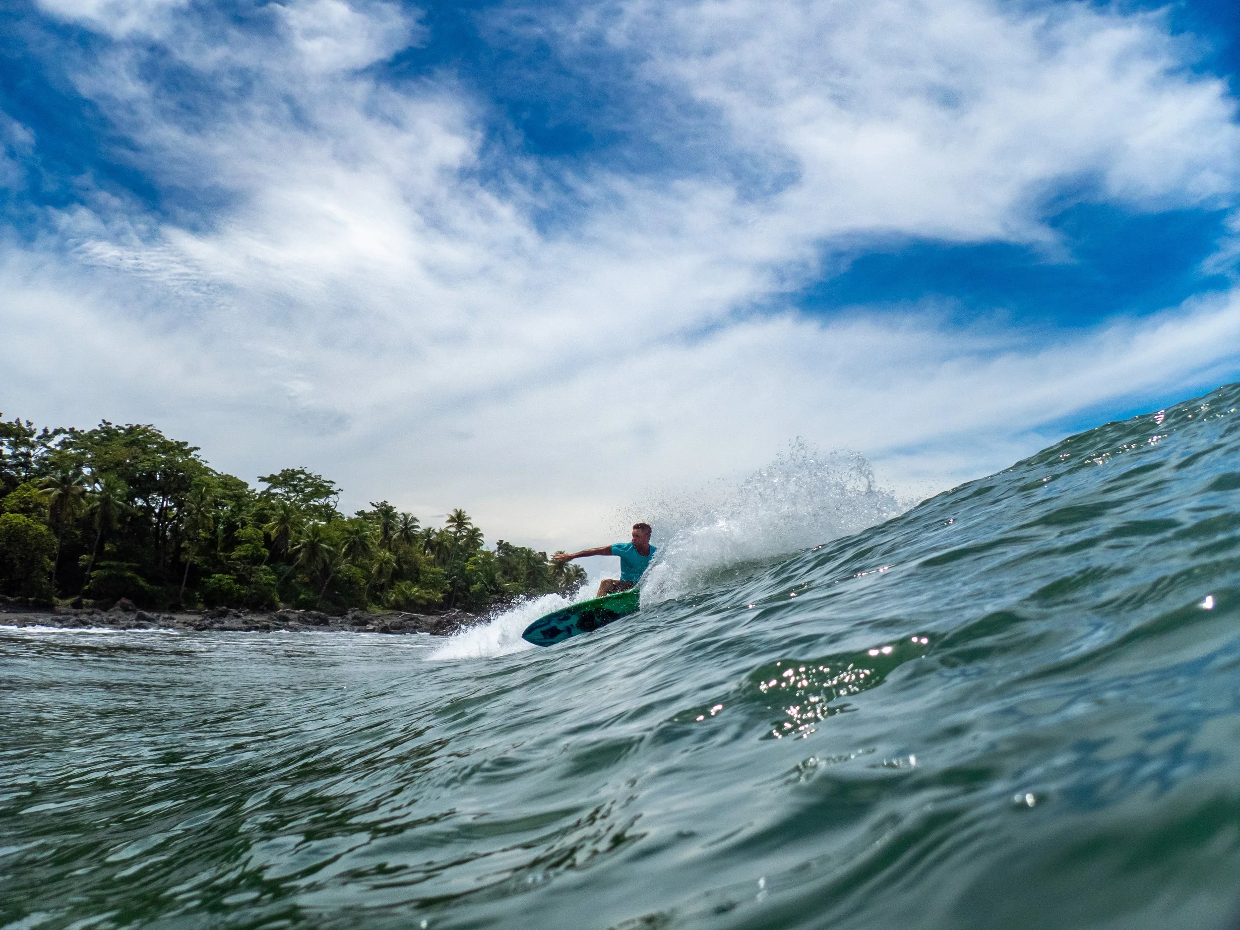 Surfer riding a wave near a lush, tropical coastline with a blue sky and scattered clouds.