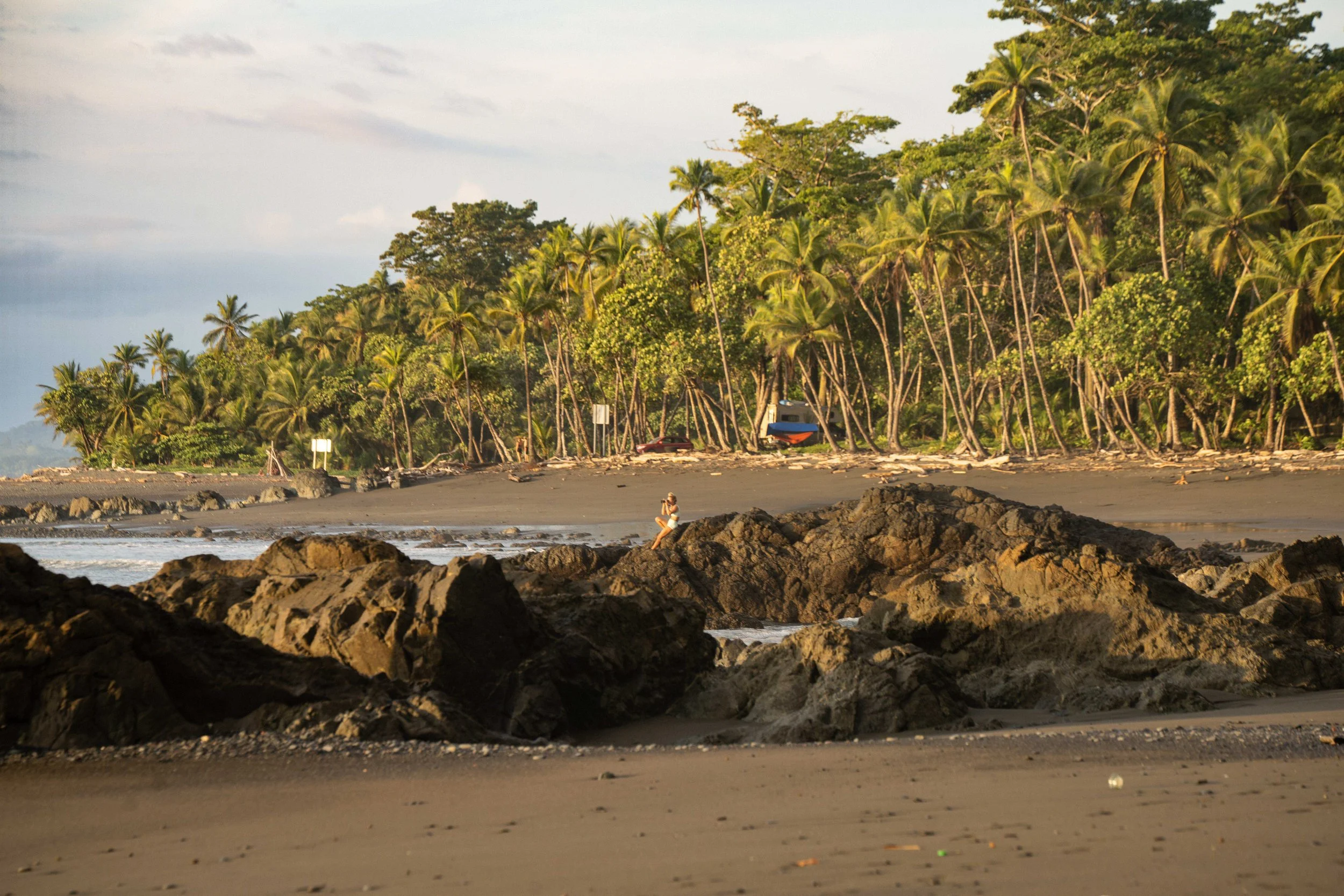 A scenic beach with rocks in the foreground, a woman sitting on a large rock, and a lush backdrop of palm trees and other greenery under a cloudy sky.