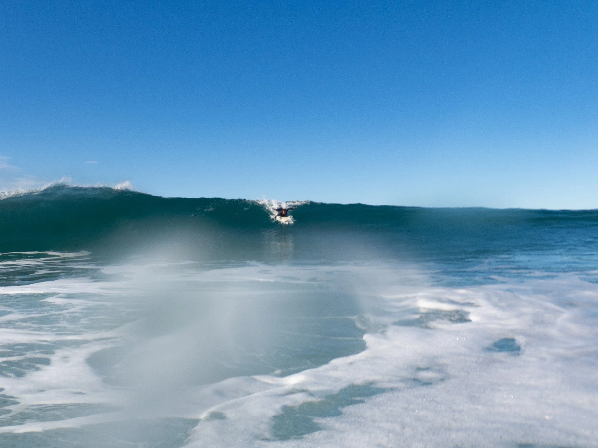 A surfer riding a large wave on the open ocean under a clear blue sky.