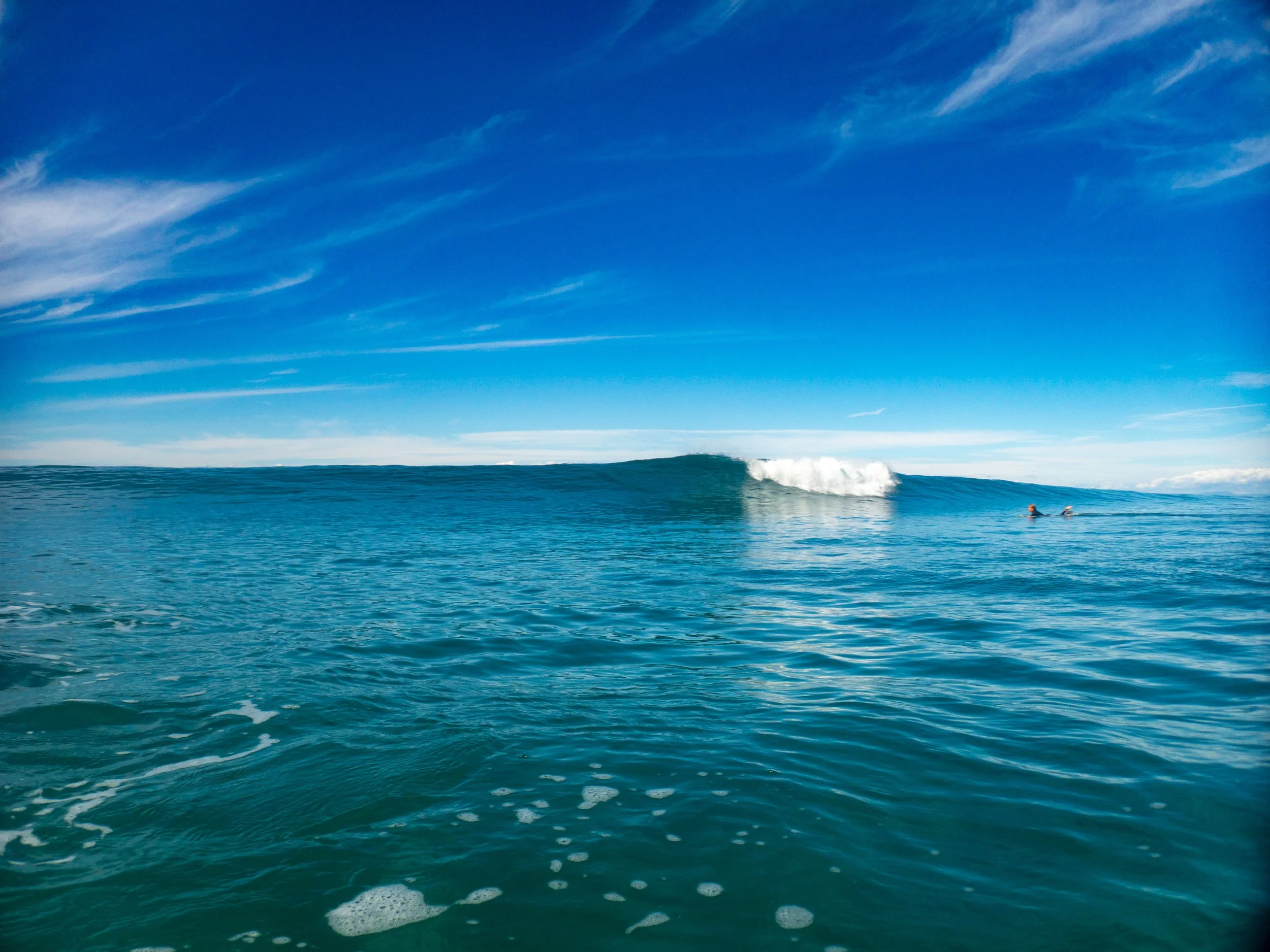 Surfer paddling on a surfboard towards a large wave in an open ocean under a clear blue sky with a few wispy clouds.