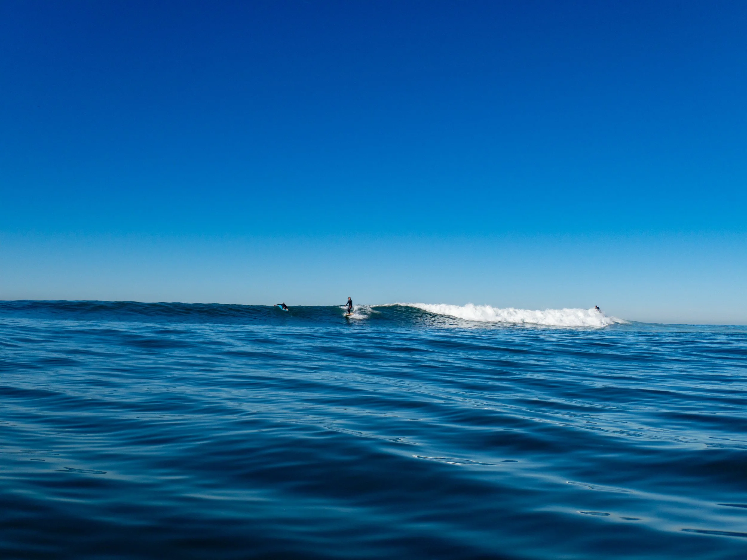 Three surfers riding and paddling on ocean waves, with a clear blue sky above.